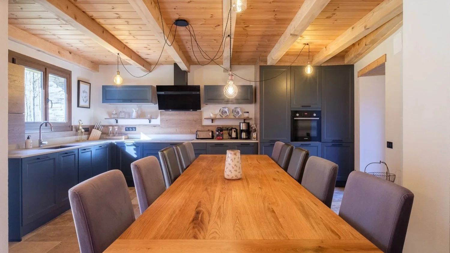 Cuisine moderne avec plan de travail blanc, armoires grises, table en bois, chaises grises, lumière suspendue, fenêtre, et murs en bois.