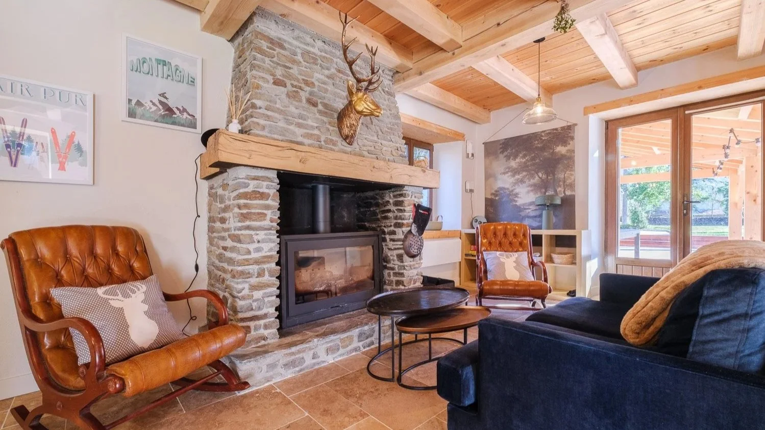 Salon avec cheminée en pierre, végétation artificielle, meubles en cuir et tissu, porte-fenêtre en bois laissant entrer la lumière naturelle, décor rustique.