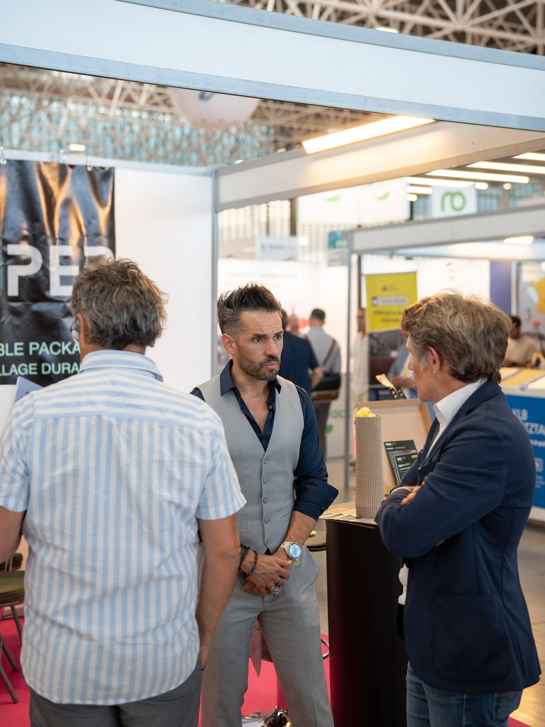 Trois hommes travaillent et discutent dans un stand d'exposition à une foire ou un salon professionnel, avec un panneau d'affichage derrière eux.