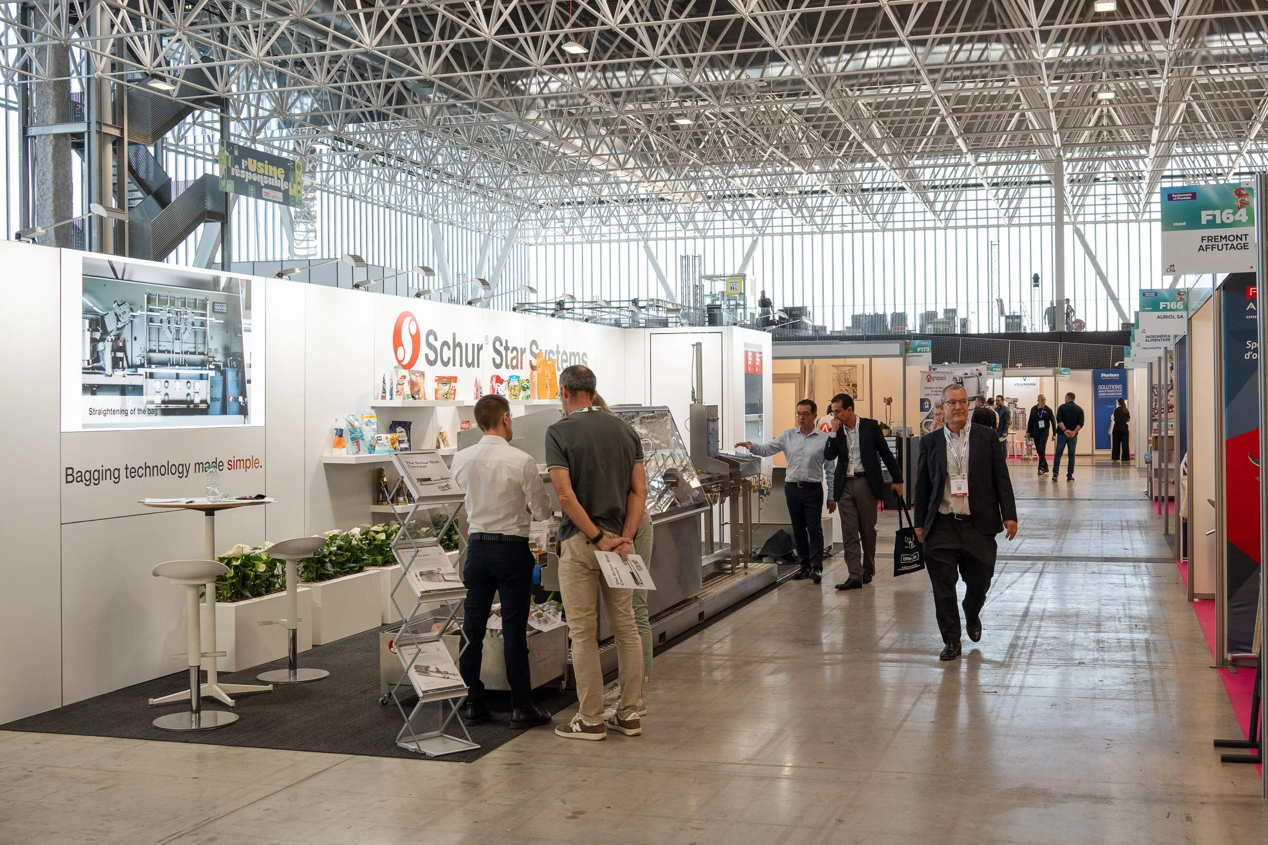Vue intérieure d'un hall d'aéroport avec plusieurs personnes discutant et se déplaçant, des stands d'exposition liés à la technologie, un plafond de structure métallique, et une grande fenêtre laissant entrer la lumière naturelle.