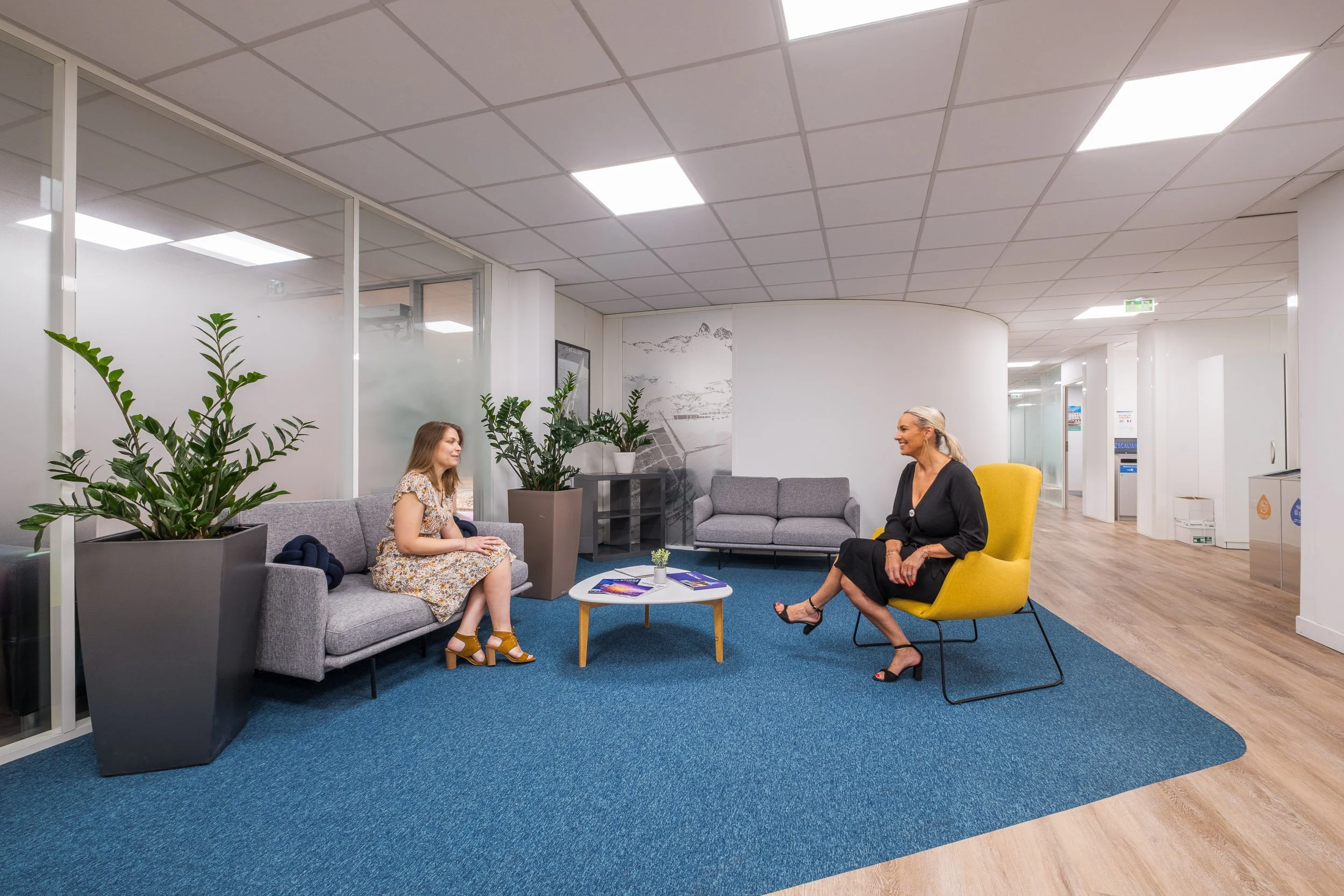 Deux femmes discutent dans un espace d'attente moderne avec des canapés gris, une table ronde, des plantes vertes en pot et un mur décoré avec une peinture de montagnes et un quai.