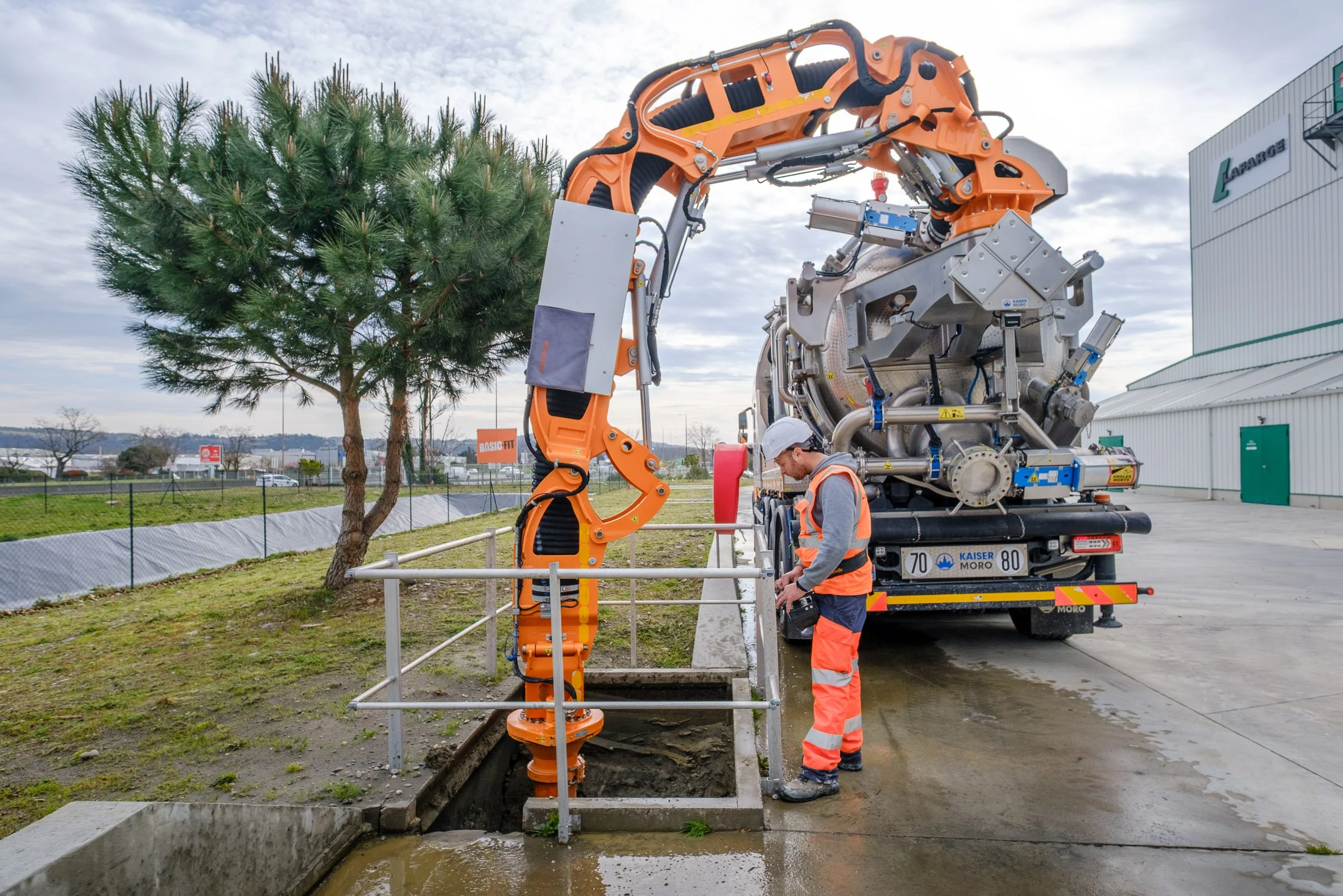 Travailleur en sécurité utilisant une machine pour réparer une canalisation d'eau souterraine. Il porte un casque blanc et un gilet de sécurité orange. La machine orange est attachée à un camion dans une zone extérieure.