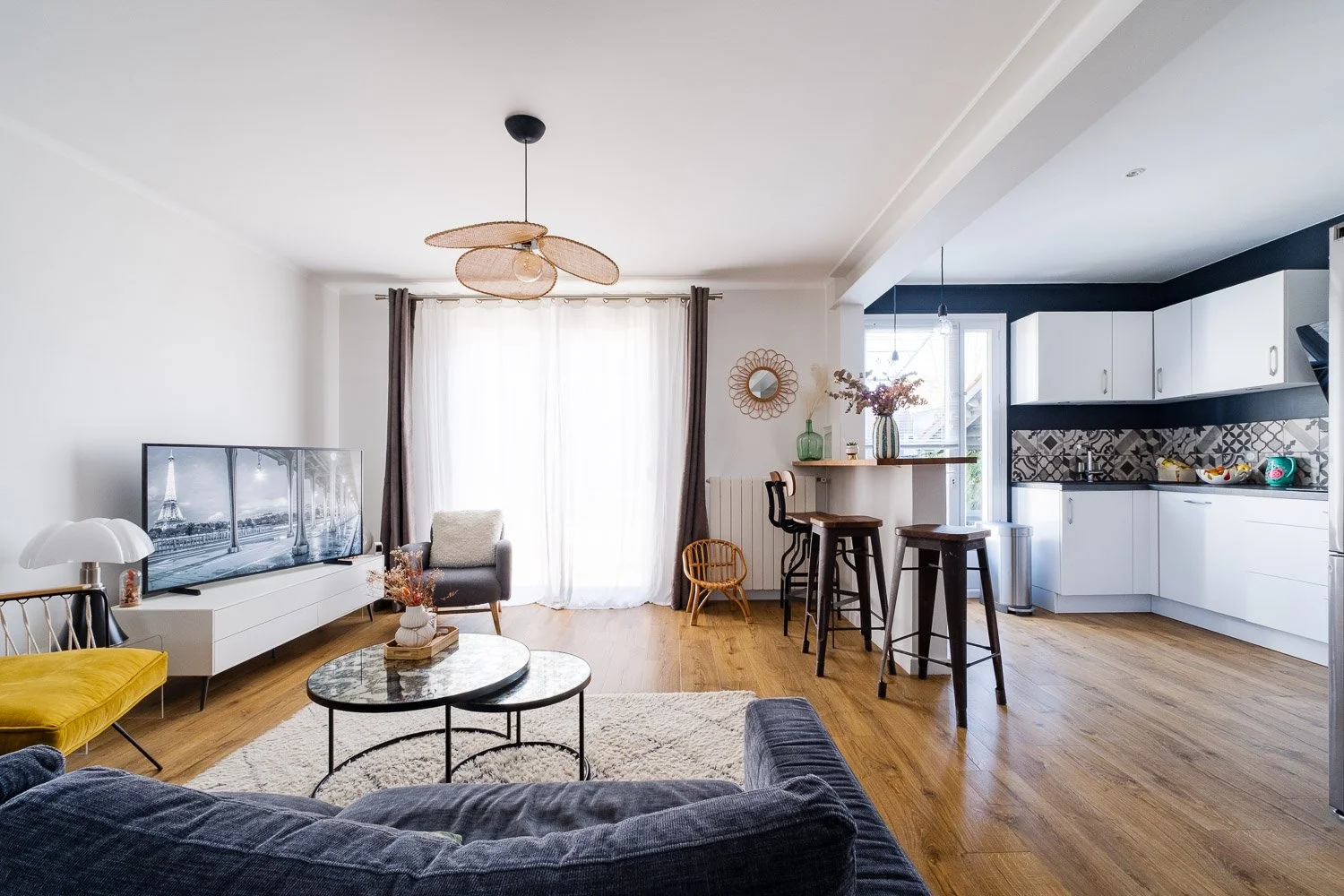 Salon et cuisine ouverts avec meubles modernes, télévision, canapé gris, table basse, bar avec tabourets, cuisine blanche avec crédence géométrique, décoration florale, grande fenêtre avec rideaux et sol en bois.