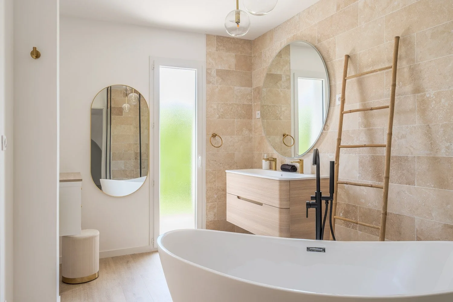 Salle de bain moderne avec baignoire autoportante, miroir rond, lavabo en bois clair, grande fenêtre, escalier en bambou comme rangement, carrelage beige et décoration minimaliste.