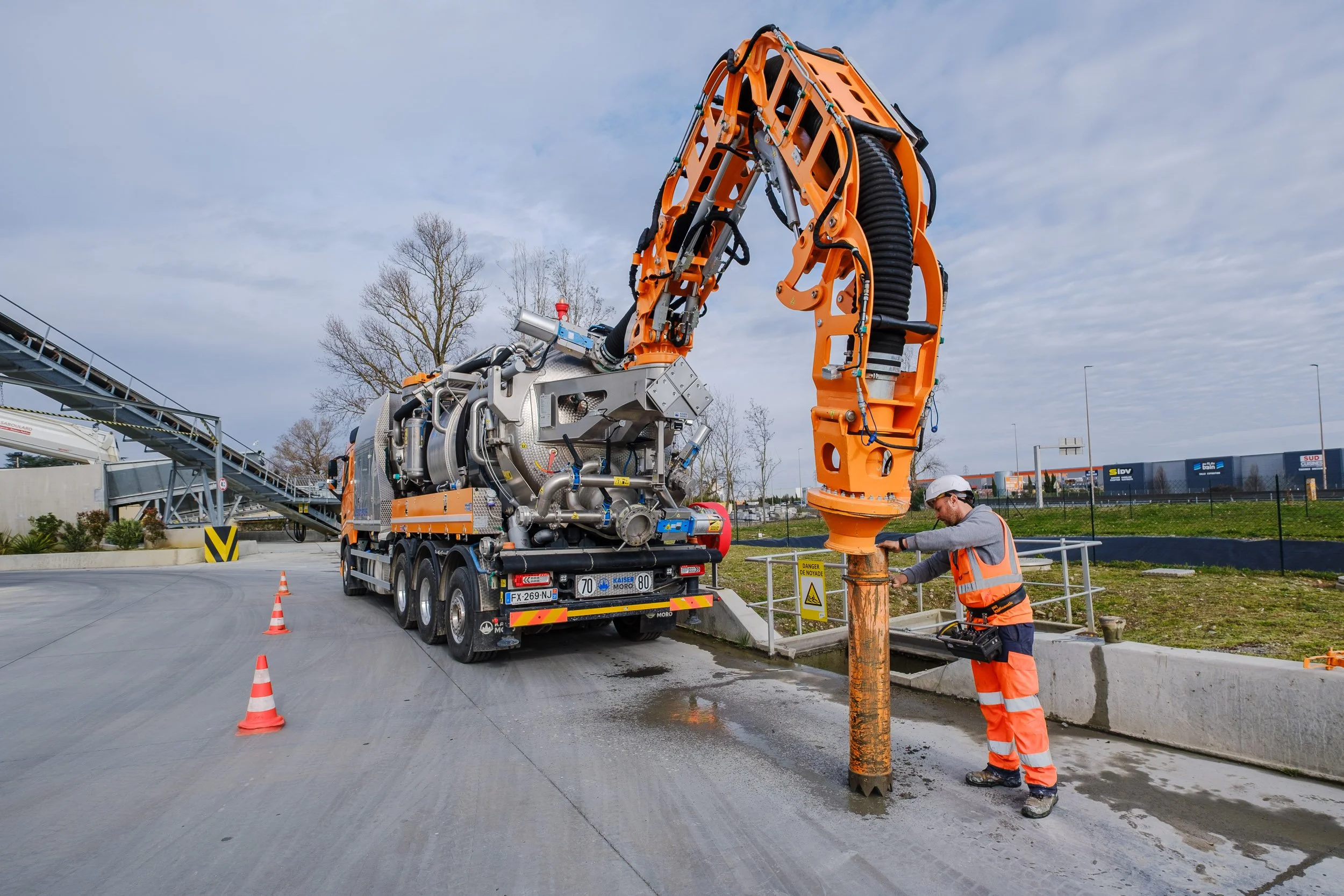 Travaux de fondation avec une machine de chantier orange et un ouvrier portant une veste et un casque de sécurité, travaillant à la pose d'une pieuvre de béton sur un parking.