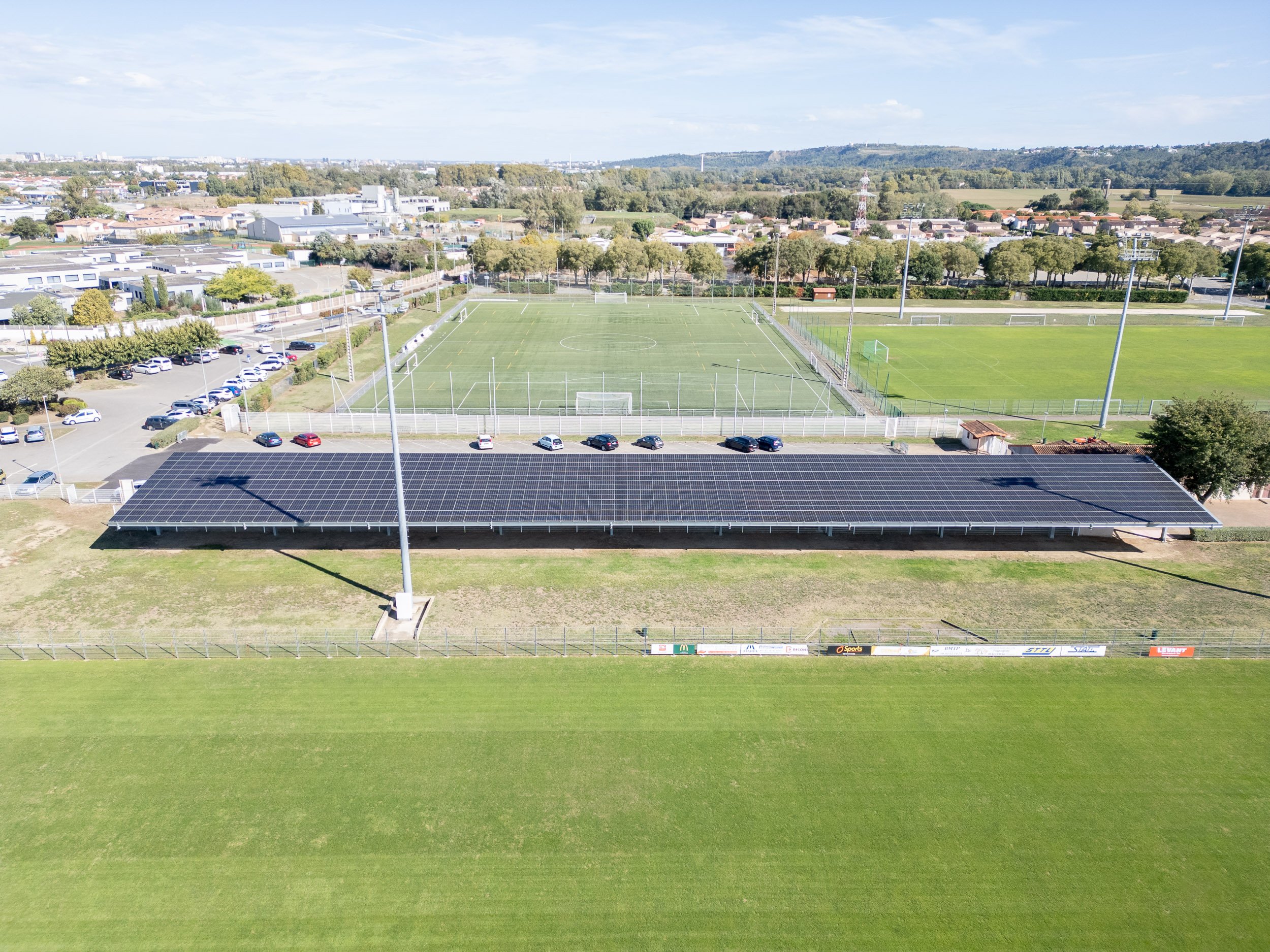 Vue aérienne d'un terrain de football avec pelouse verte, entouré de clôtures et de lampadaires, avec des installations sportives adjacentes et une zone de stationnement, dans un environnement résidentiel entouré de végétation.