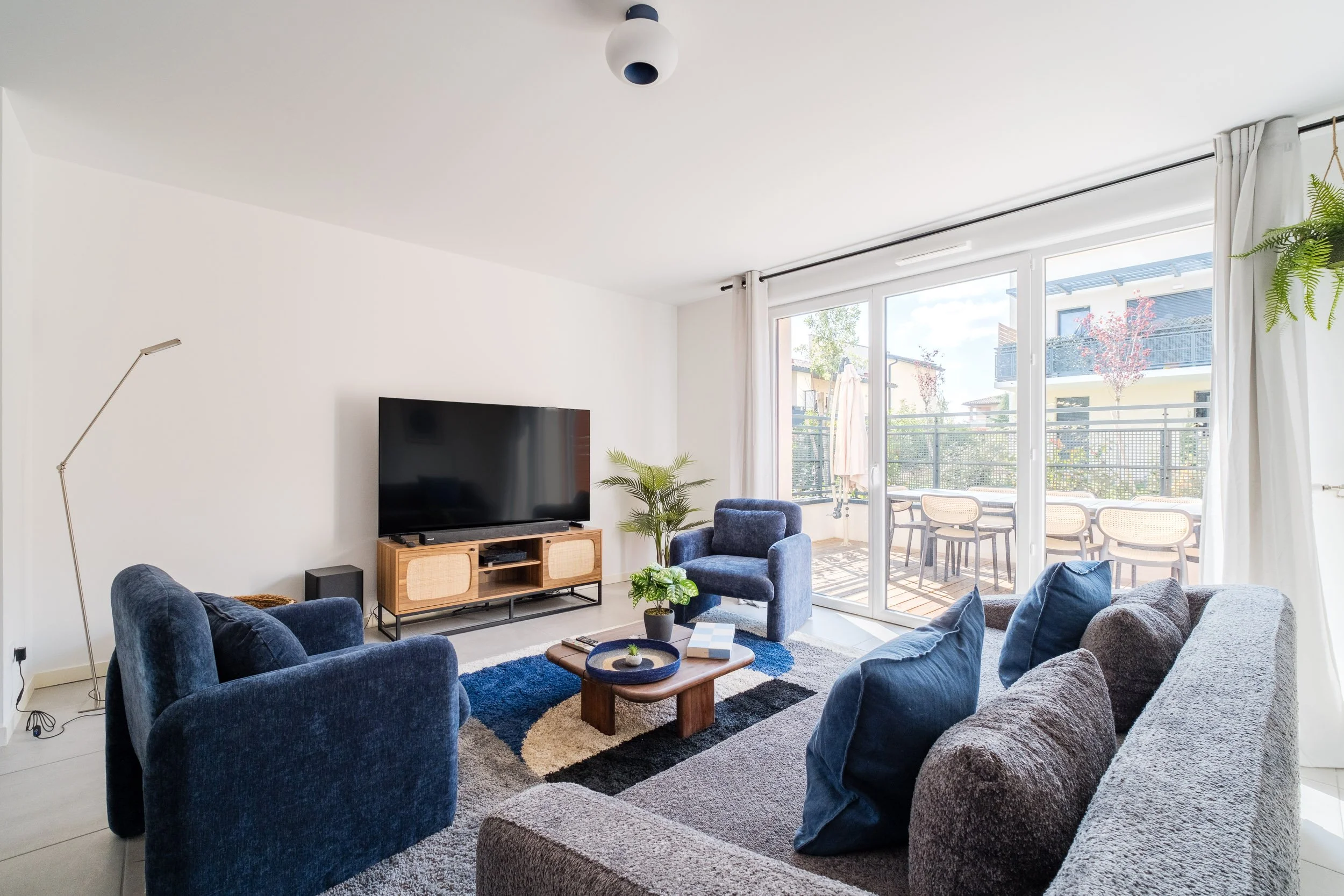 Salon moderne avec un canapé gris, des fauteuils bleu marine, une télévision sur un meuble en bois avec des étagères, une plante verte, tapis colorés, grande baie vitrée avec rideaux blancs donnant sur un balcon avec table et chaises, lumière naturel