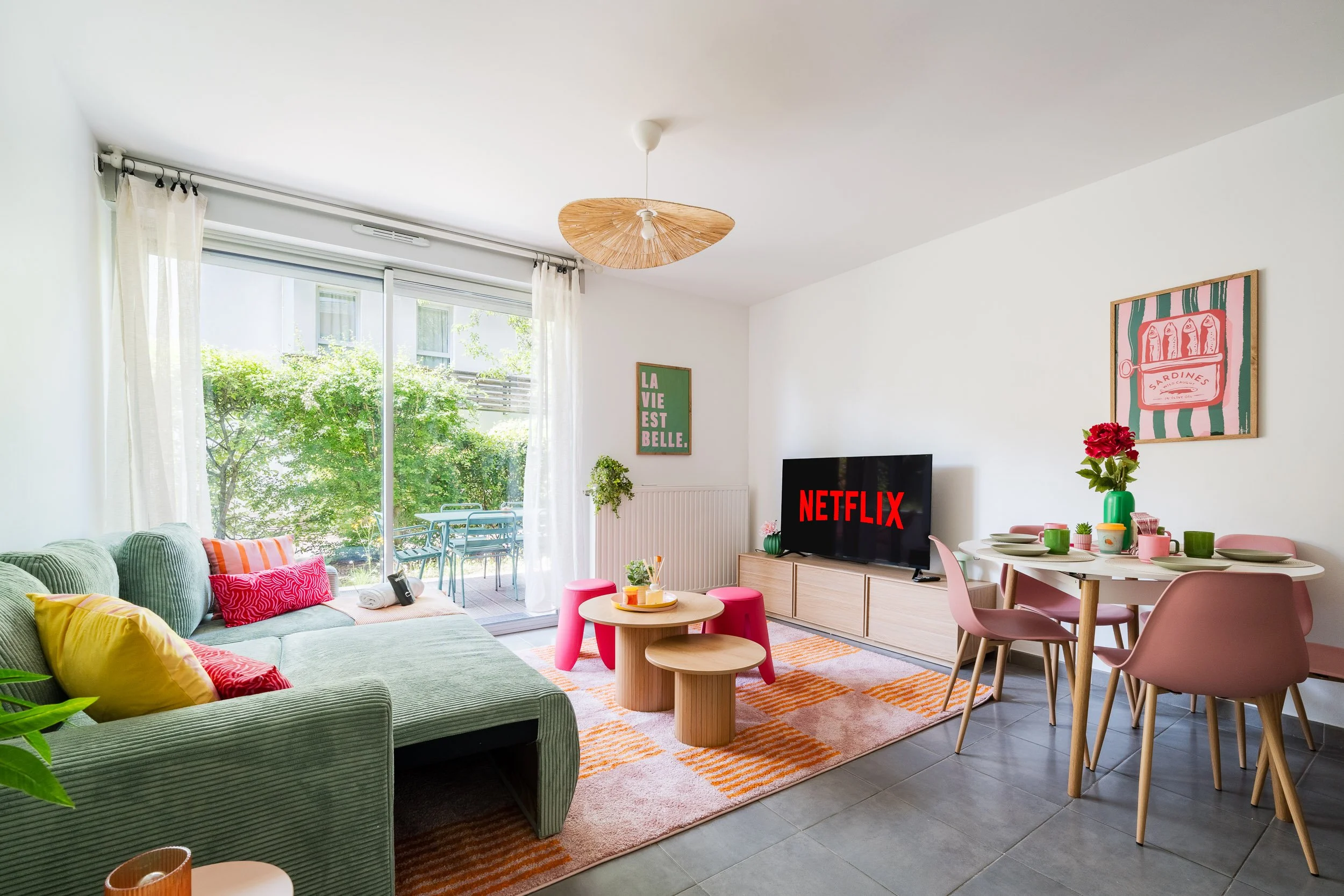 Salon lumineux avec un canapé vert, décor coloré, table basse, télévision avec logo Netflix, salle à manger avec chaises roses, fenêtres avec vue sur jardin
