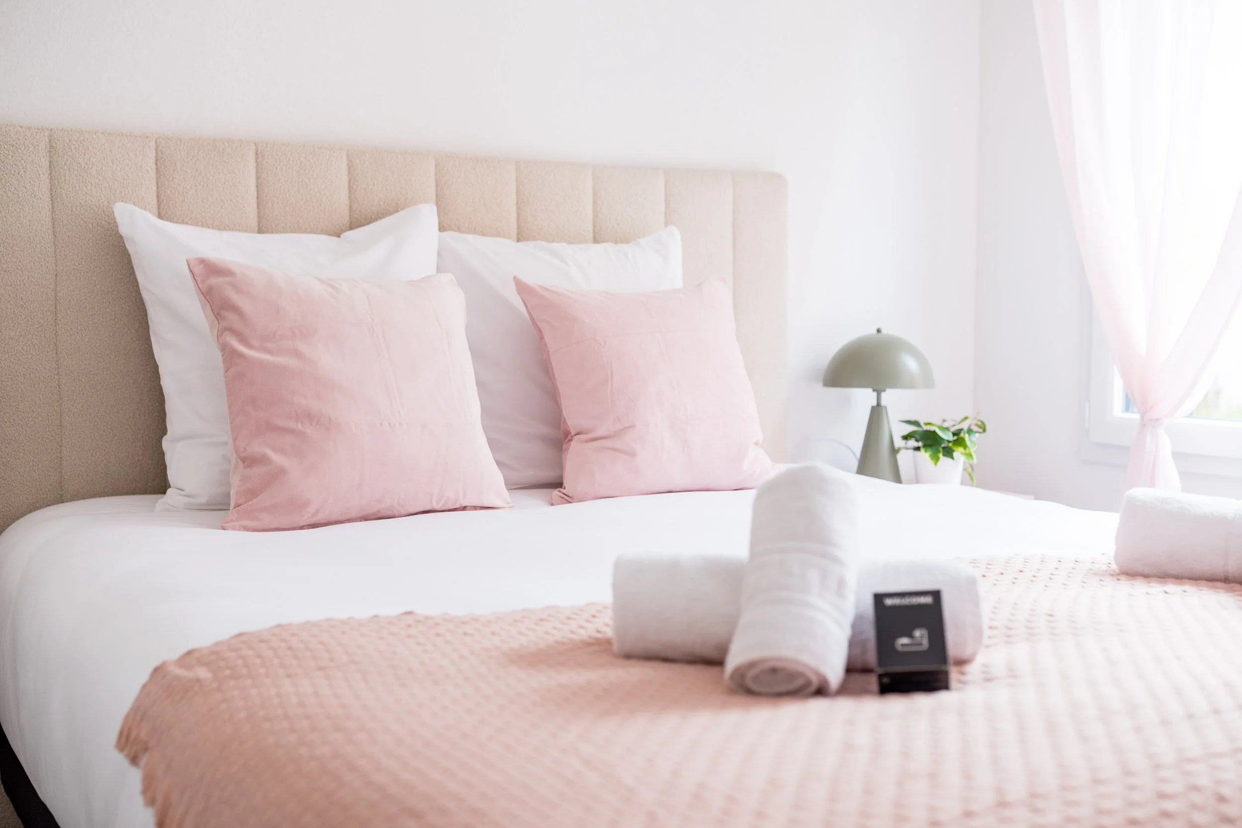 Linge de lit avec oreillers roses et blancs, serviettes blanches roulées, lampadaire, plante verte, fenêtre avec rideaux blancs dans une chambre