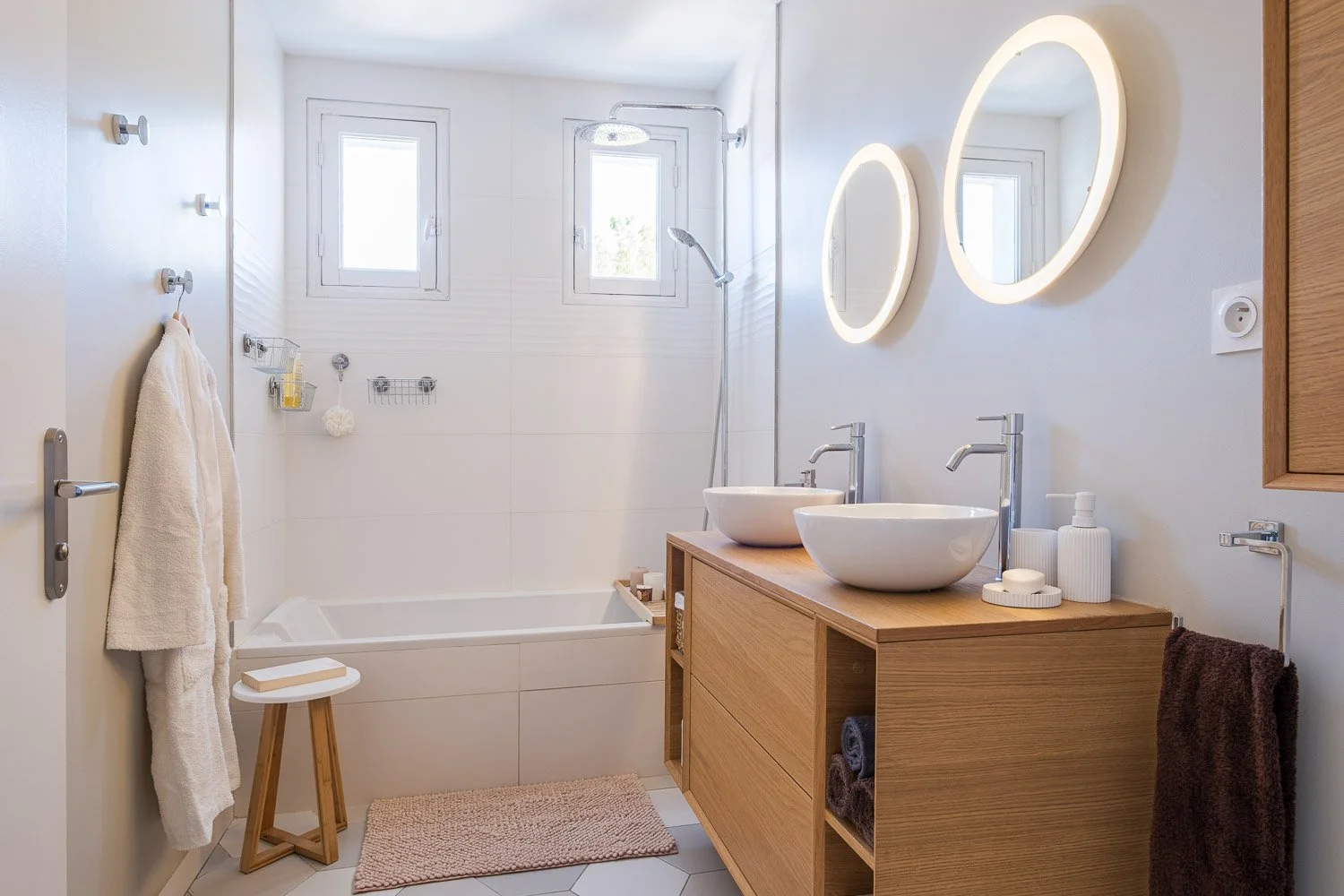 Salle de bain moderne avec double lavabo, miroir lumineux, baignoire, douches, accessoires en bois, serviettes, et petit tapis beige.