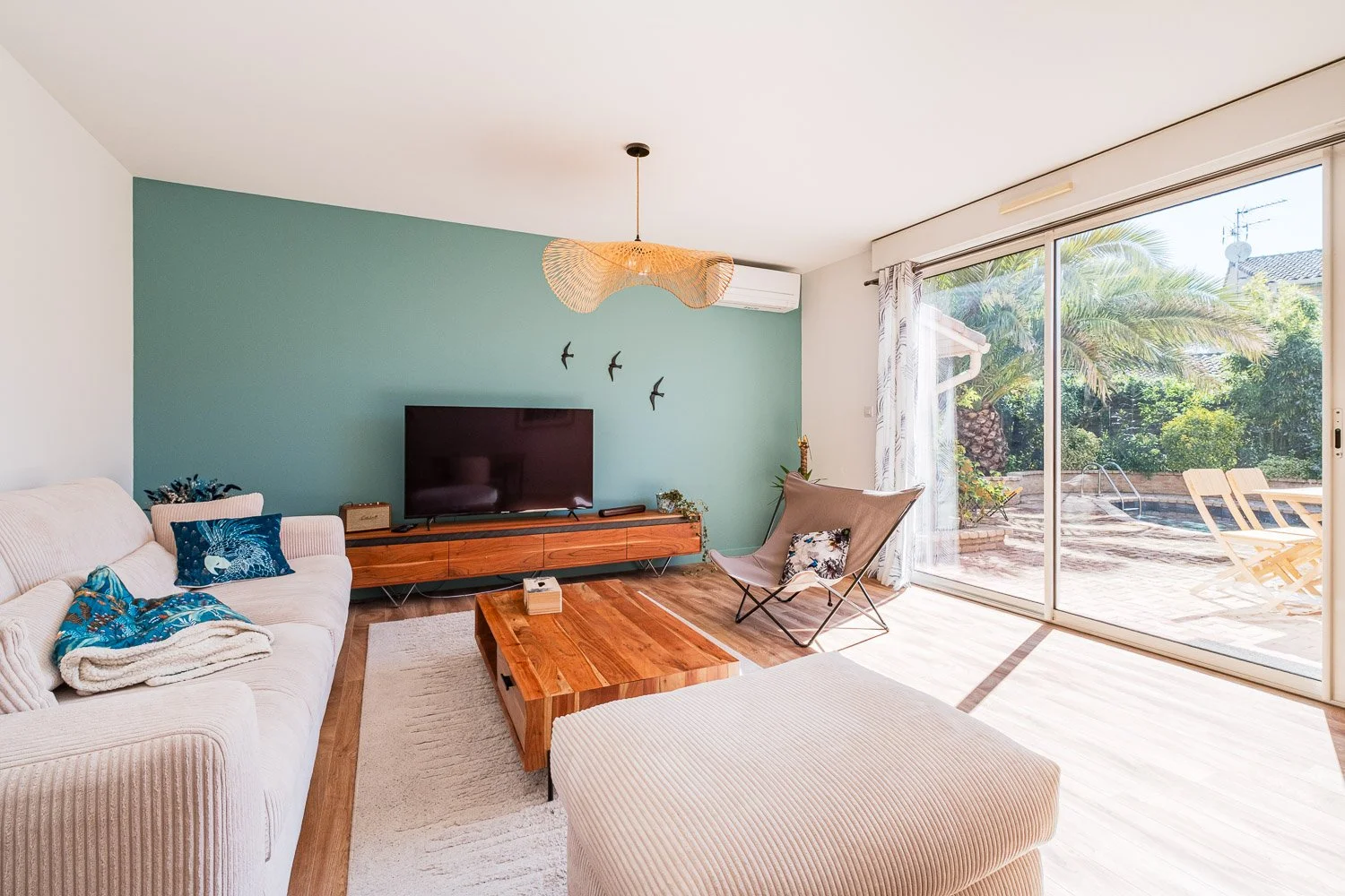 Salon lumineux avec canapé beige, téléviseur sur un meuble en bois, mur vert et grande baie vitrée donnant sur un espace extérieur avec piscine et palmier.