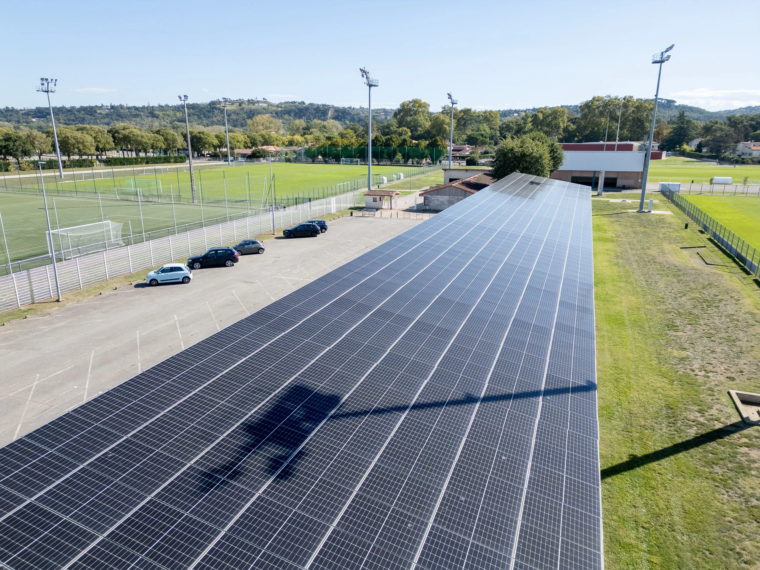Un terrain de sport avec des lampadaires, des voitures dans un parking, des terrains de sports et des arbres au fond, avec un bâtiment doté de panneaux solaires sur le toit.