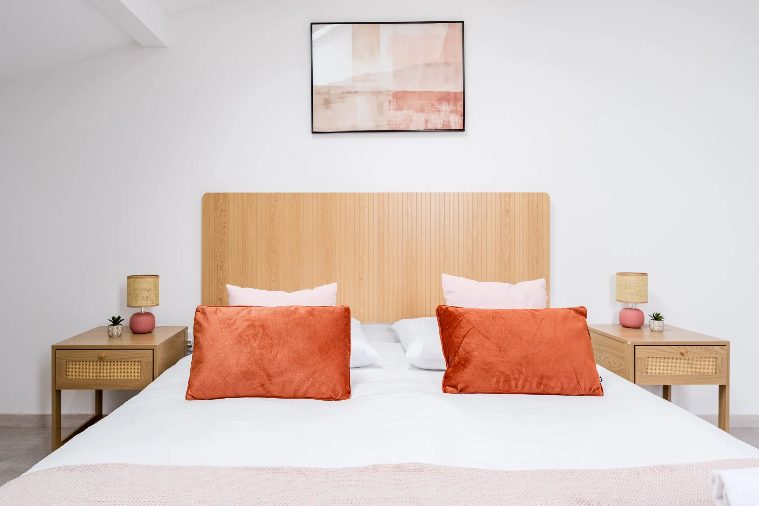 Chambre minimaliste avec lit à baldaquin blanc, oreillers blancs et coussins en velours orange, deux tables de nuit en bois clair, deux lampes en céramique rose, une peinture abstraite dans des tons de beige et de rose, murs blancs, sol en bois clair.