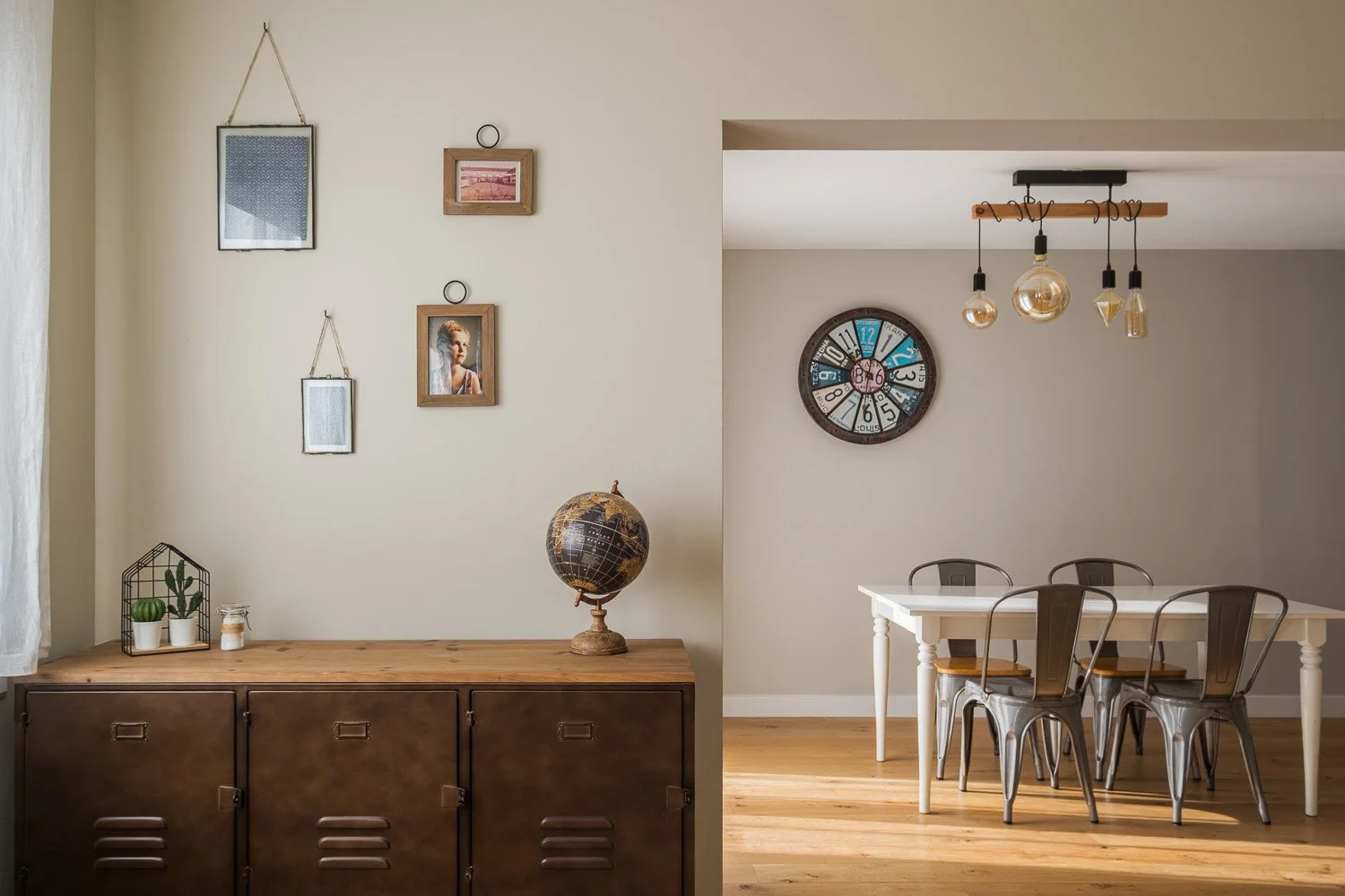 Une salle à manger avec une table blanche, six chaises en métal, un mur avec un grand horloge ronde colorée, un plafonnier avec plusieurs ampoules suspendues, et un mur crème avec des cadres photo et un globe sur un meuble en bois.