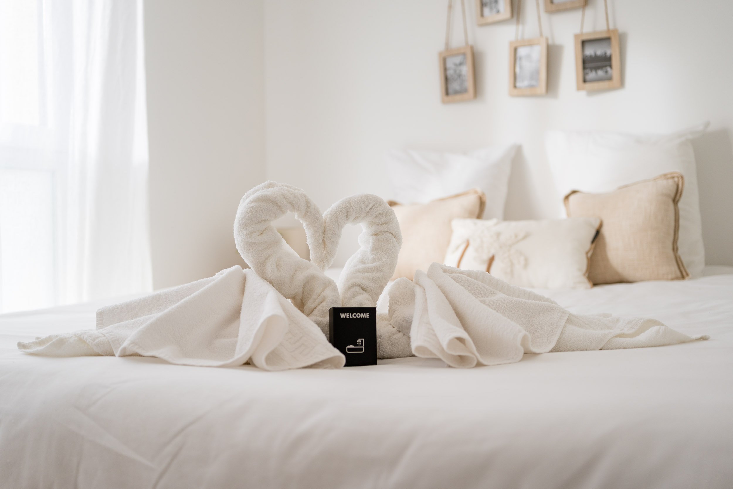 L'image montre un lit blanc avec deux serviettes en forme de cœurs, un petit panneau 'Welcome' et une prise électrique à côté. Le décor est simple et lumineux, avec des cadres photo noirs et blancs suspendus au mur à l'arrière.