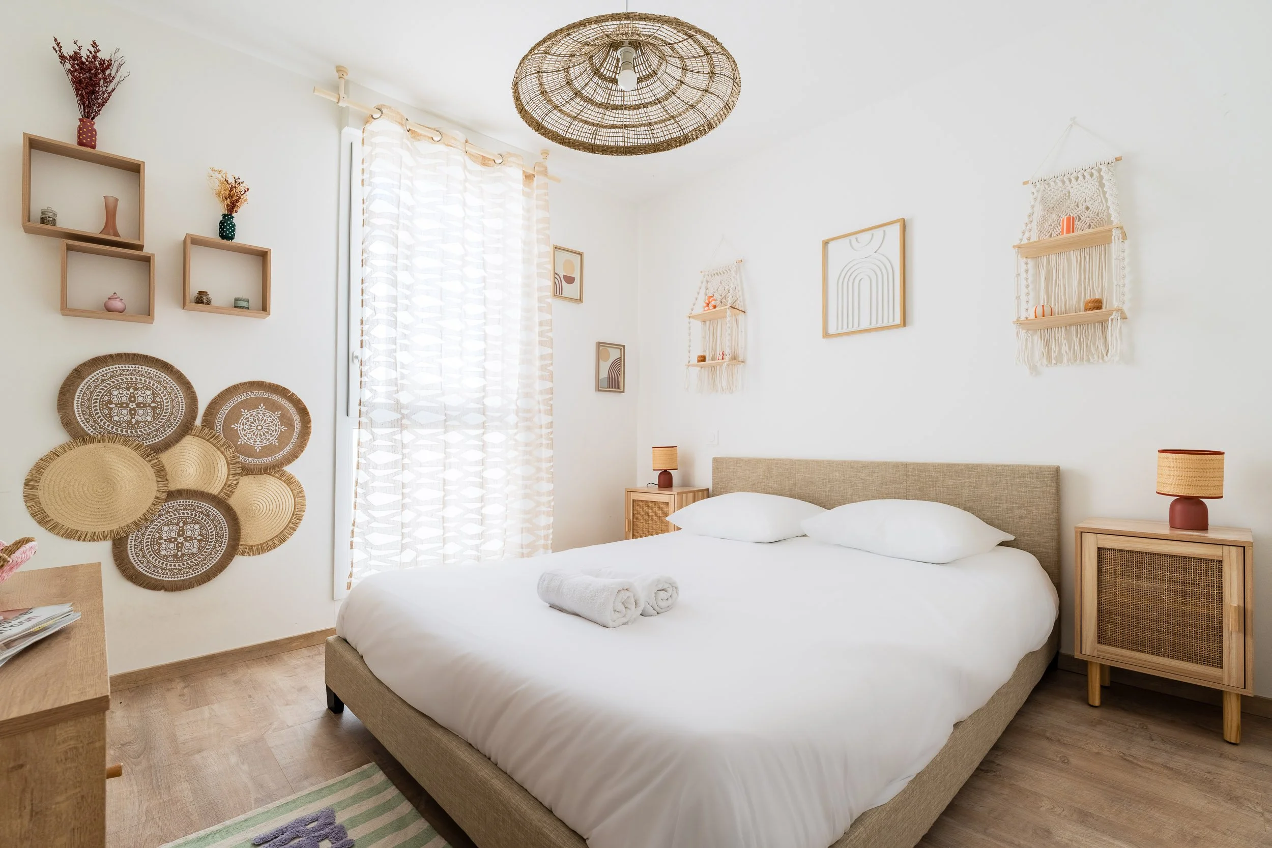 Chambre décorée dans un style bohème avec un lit double, plusieurs cadres muraux, des étagères en bois avec des objets décoratifs, un plafond en rotin, et des rideaux légers.