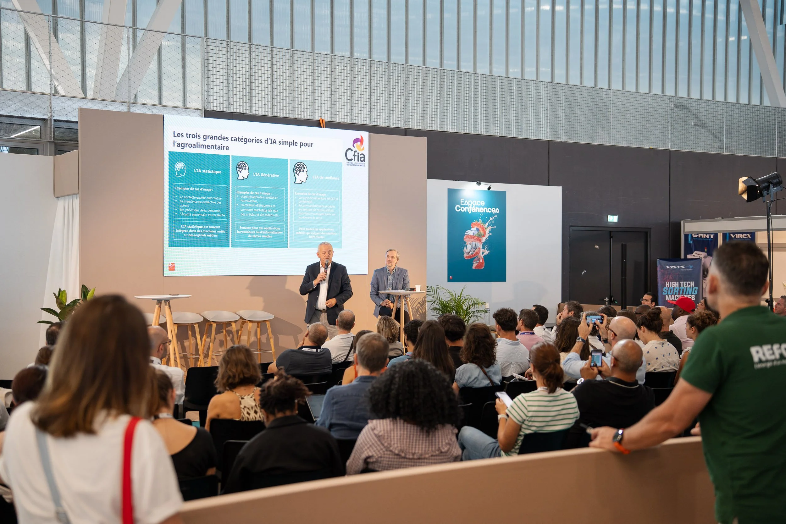 Une conférence dans un grand espace intérieur avec un public attentif. Sur scène, deux hommes parlent, l'un tient un micro. Un grand écran affiche une présentation sur les catégories de l'intelligence artificielle dans l'alimentaire. La salle est mod