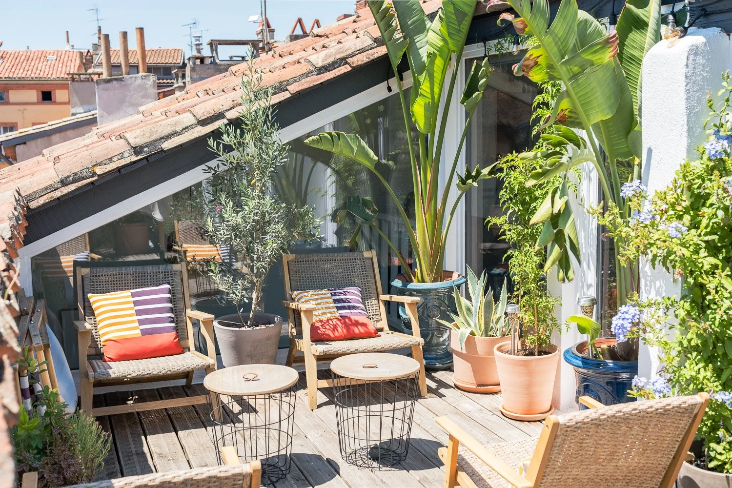 Balcon ensoleillé avec des fauteuils en rotin, des coussins rayés, et plusieurs pots de plantes vertes et fleuries.