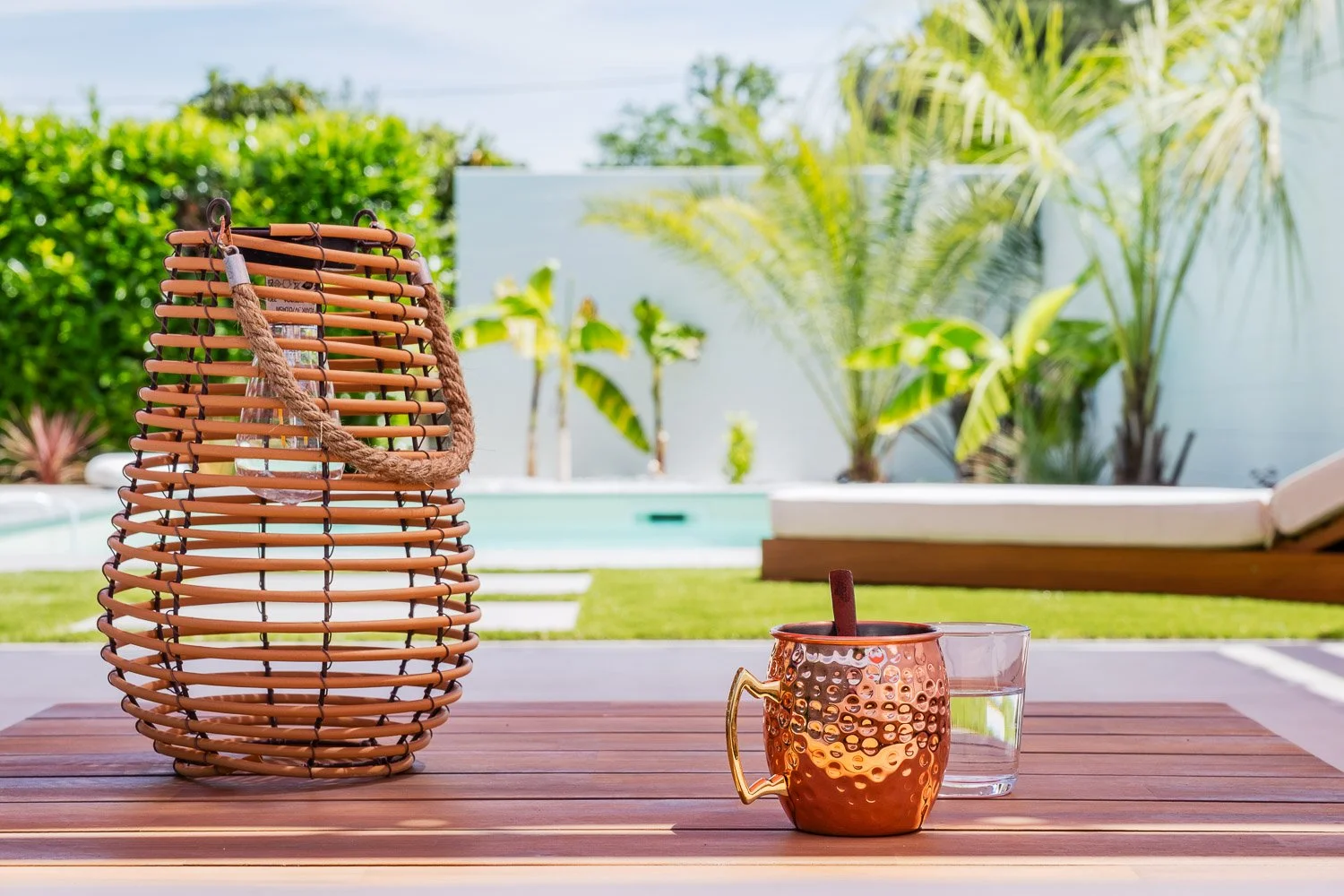 Une table en bois avec une carafe en rotin contenant de l'eau, un verre vide, et une tasse en cuivre avec une cuillère en bois, à l'extérieur dans un jardin avec du mobilier et des plantes tropicales.