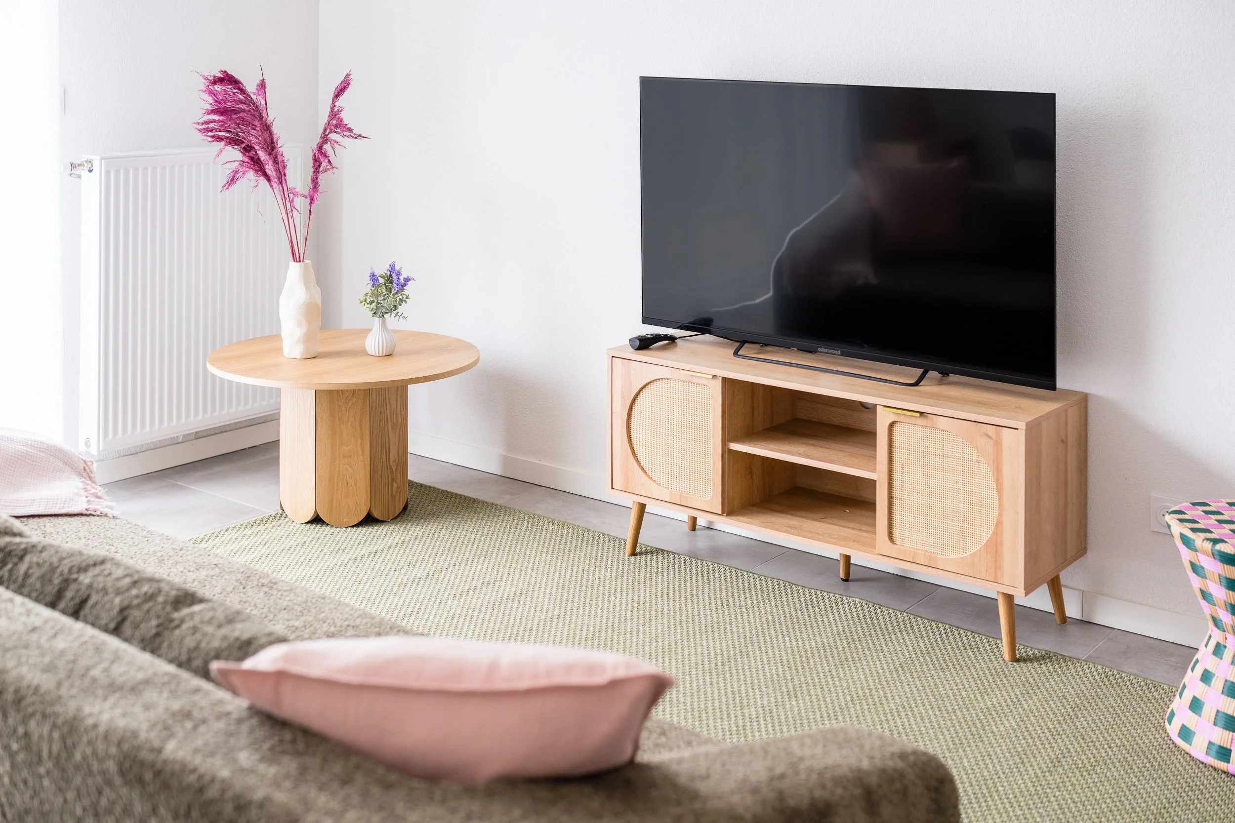 Un salon moderne avec un meuble en bois, une grande télévision, et deux vases avec des fleurs, un tapis vert sur le sol.