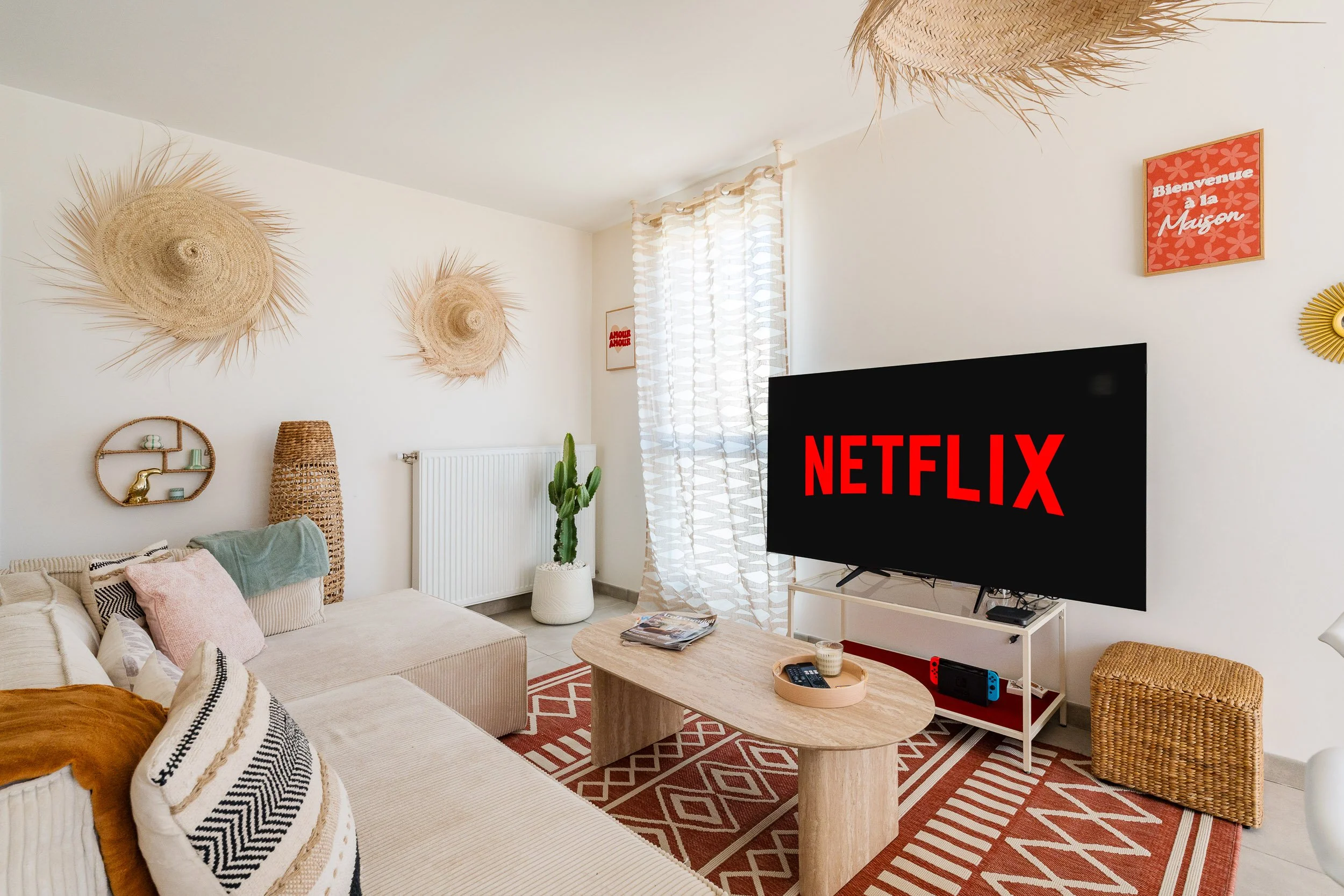 Salon avec un canapé beige, un tapis rouge et blanc, une table en bois, une télévision affichant Netflix, décorations murales en fibre naturelle, un cactus, et une fenêtre avec des rideaux blancs à motifs. Mur blanc avec affiches et décorations