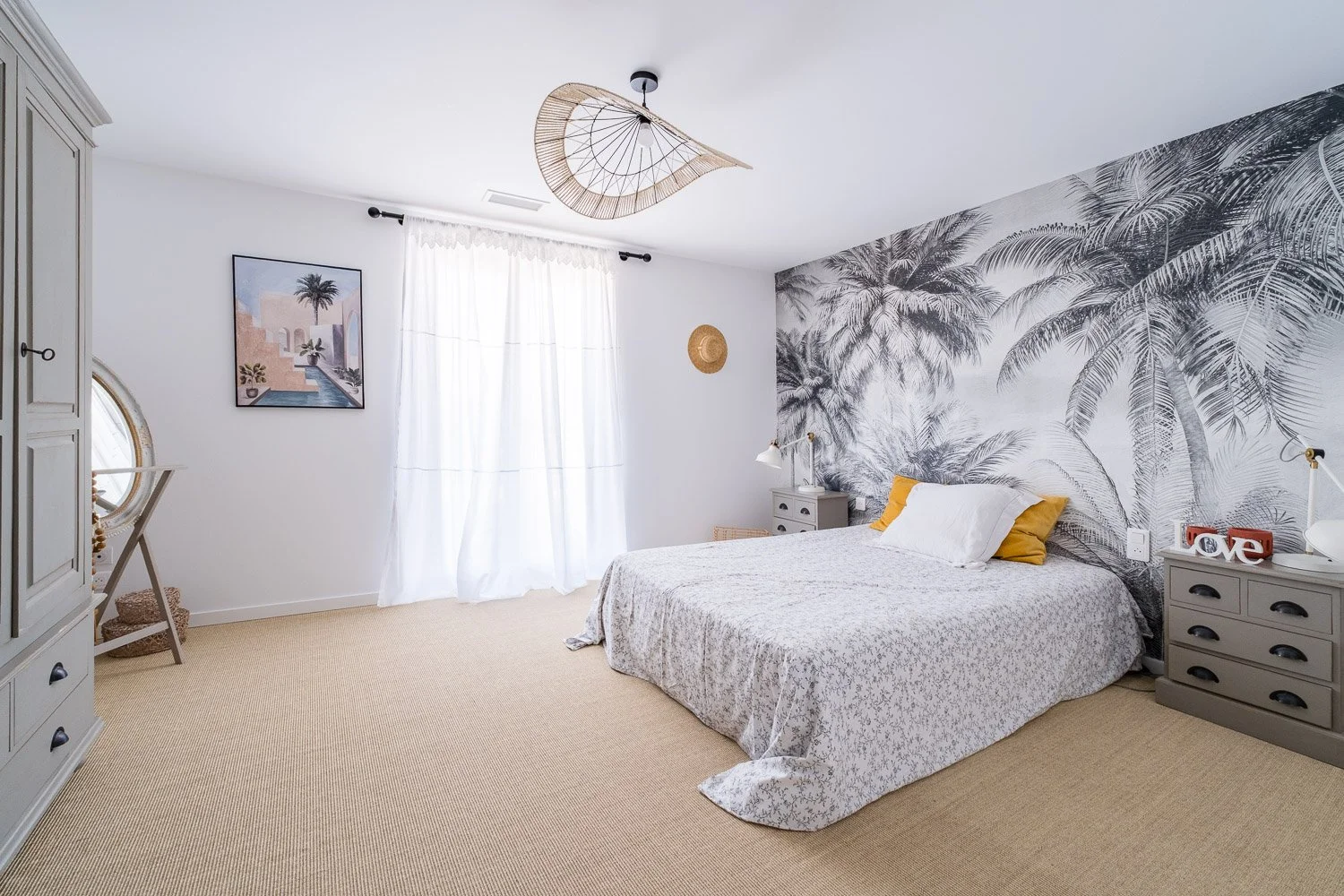 Chambre à coucher avec une tête de lit ornée d'un papier peint noir et blanc avec des palmiers. Lit avec draps blancs, oreillers jaunes et blancs. Commode grise à côté du lit, décorée d'objets comme une lampe, un cadre "love" et une montre. Fenêtre a