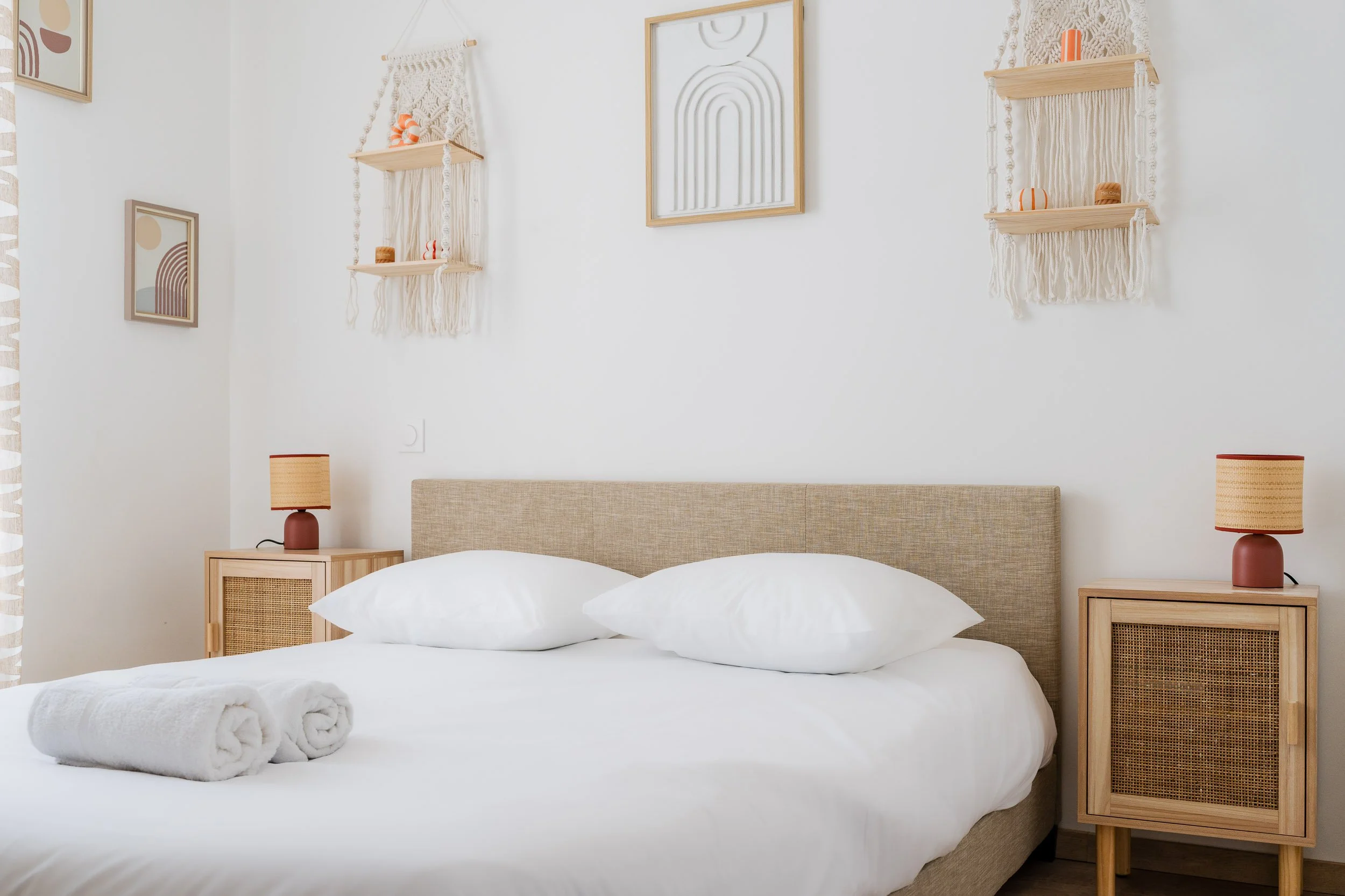 Un lit double avec un cadre en bois clair, deux oreillers blancs, deux serviettes pliées enroulées à la base du lit, deux tables de chevet en bois clair avec des lampes orange à côté, murs blancs avec décorations en macramé, cadres d'art abstrait, et