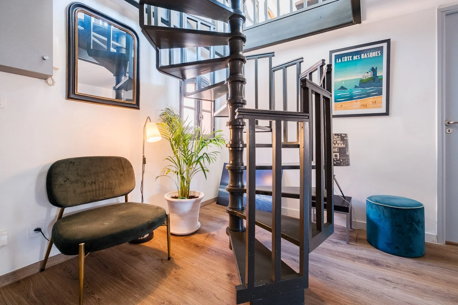 Salon intérieur avec un escalier en bois sombre, un fauteuil vert en velours, une plante verte en pot, une lampe sur le sol, une télévision, et deux tableaux, dont un avec une scène maritime