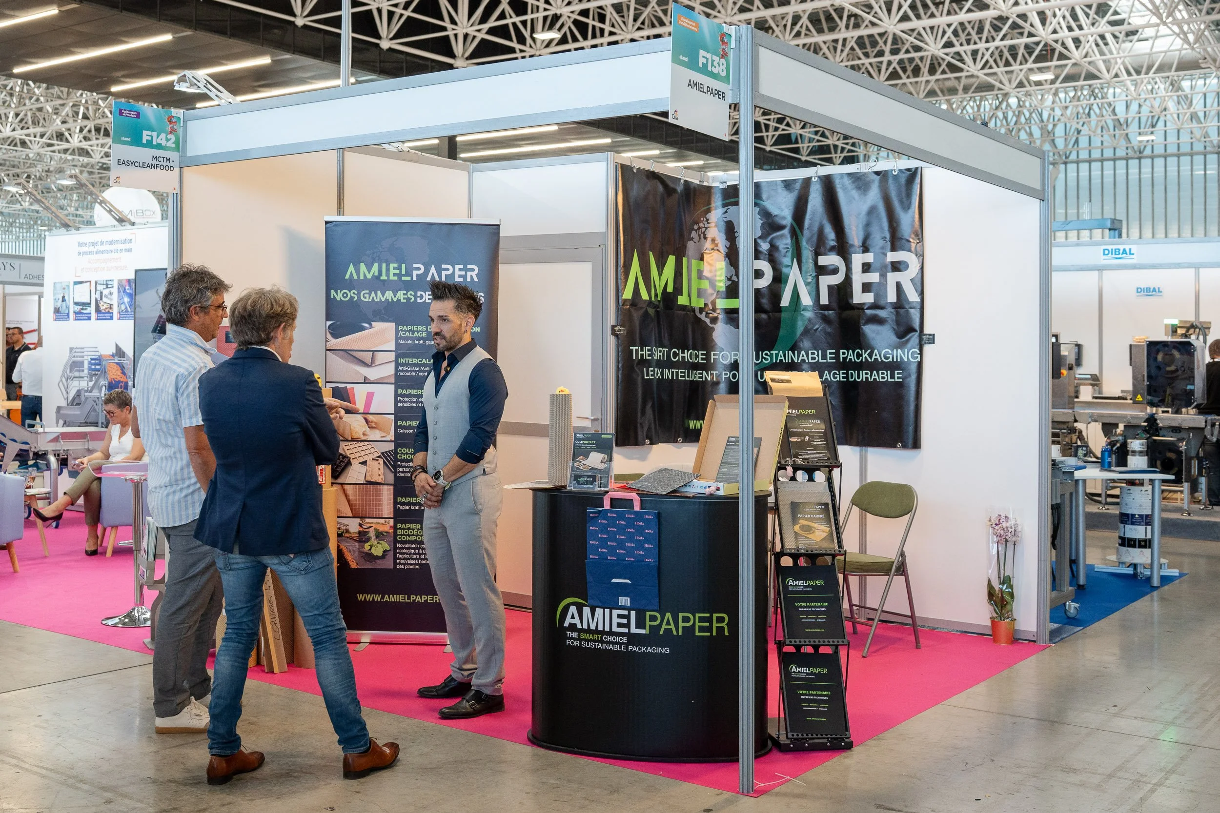 Un stand d'exposition lors d'un salon avec plusieurs personnes discutant. Le stand appartient à Amiel Paper, une entreprise spécialisée dans l'emballage durable, avec des affiches, brochures et produits exposés.