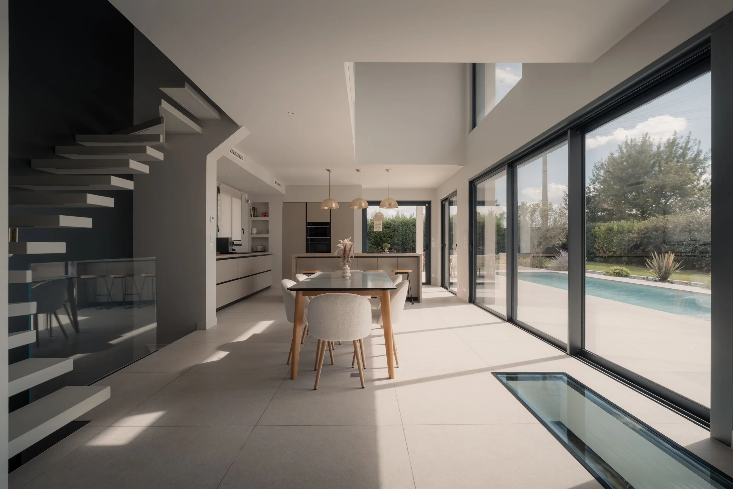 Intérieur moderne d'une maison avec grandes fenêtres laissant entrer la lumière naturelle, vue sur un jardin avec piscine extérieure, mobilier de cuisine minimaliste, et quelques éléments de décoration neutres.