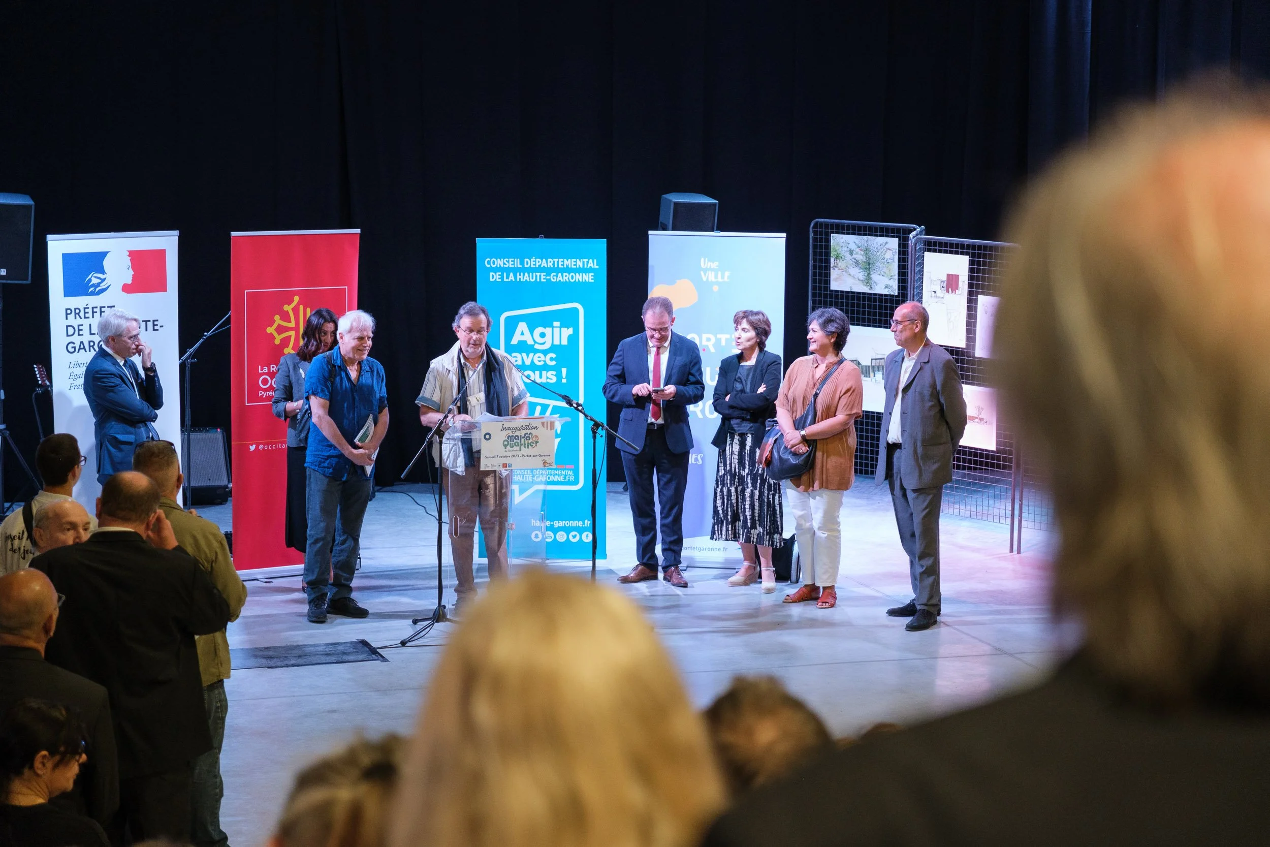 Une scène d'inauguration ou de cérémonie officielle avec plusieurs personnes sur scène, certains parlant ou regardant vers le public, entourées de banners et d'affiches promotionnelles. L'événement semble se dérouler dans un lieu intérieur avec des œ