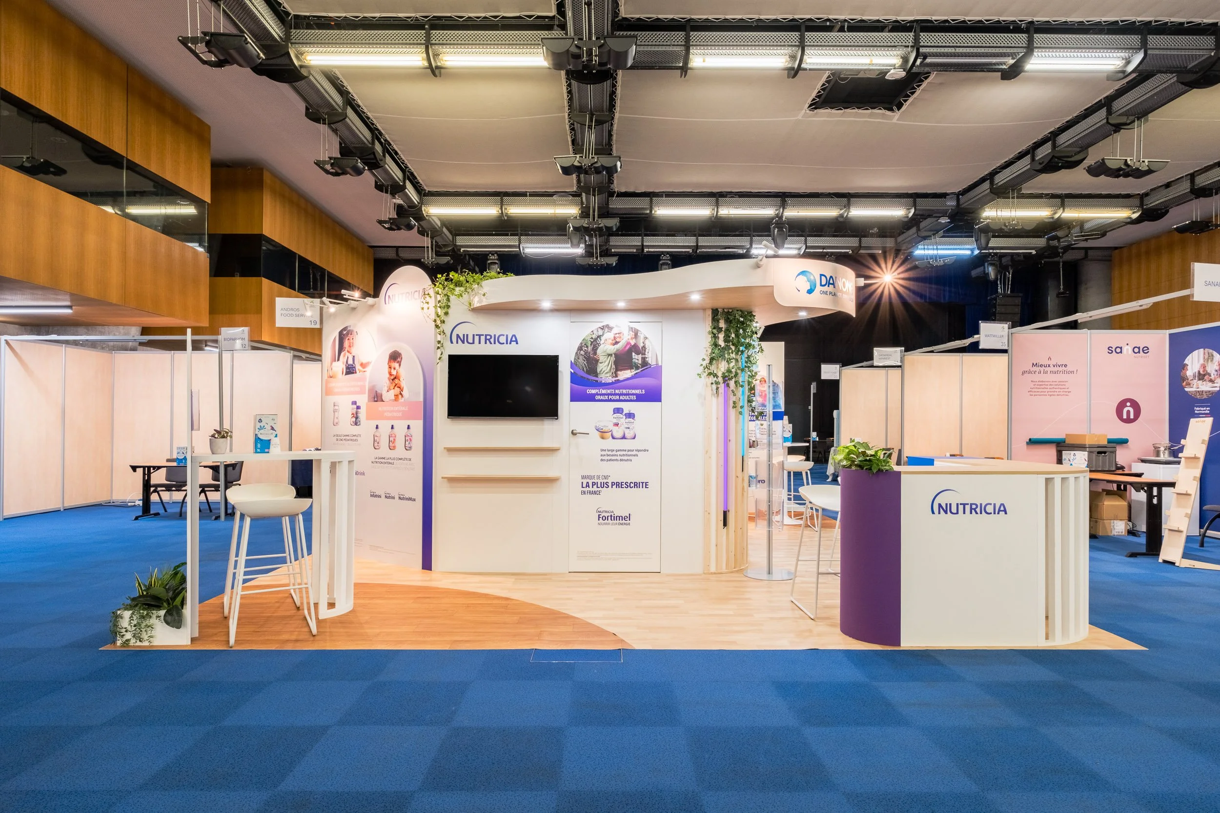 Stand d'exposition pour Nutricia avec présentoirs, panneaux informatifs, et une table avec des chaises, le tout dans un espace intérieur avec sol bleu et plafond avec éclairage suspendu.