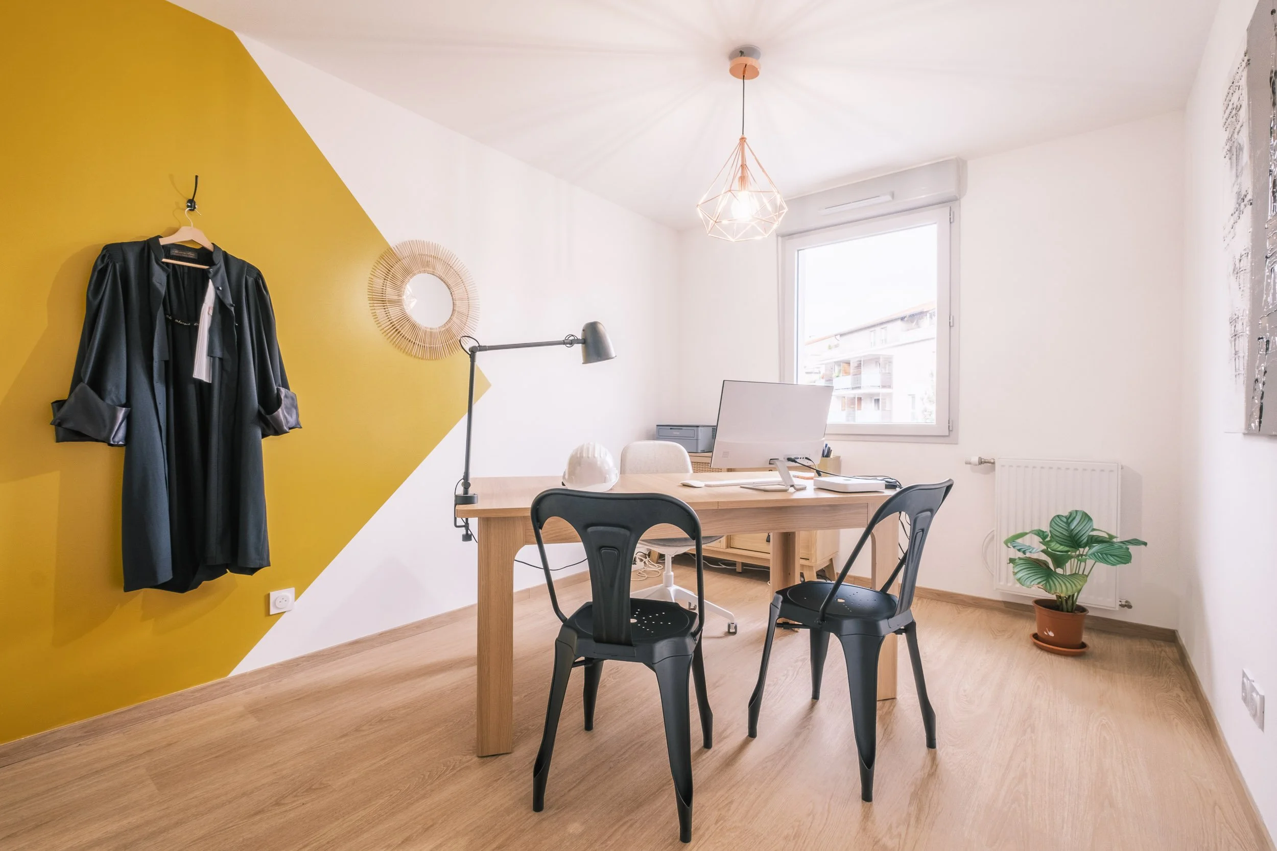 Un bureau avec une table en bois, trois chaises en plastique noires, une lampe de bureau, une plante verte en pot, une fenêtre laissant entrer la lumière naturelle, une décoration murale ronde, un vestiaire avec un manteau noir suspendu, une lampe suspendue au plafond, et un radiateur sous la fenêtre.