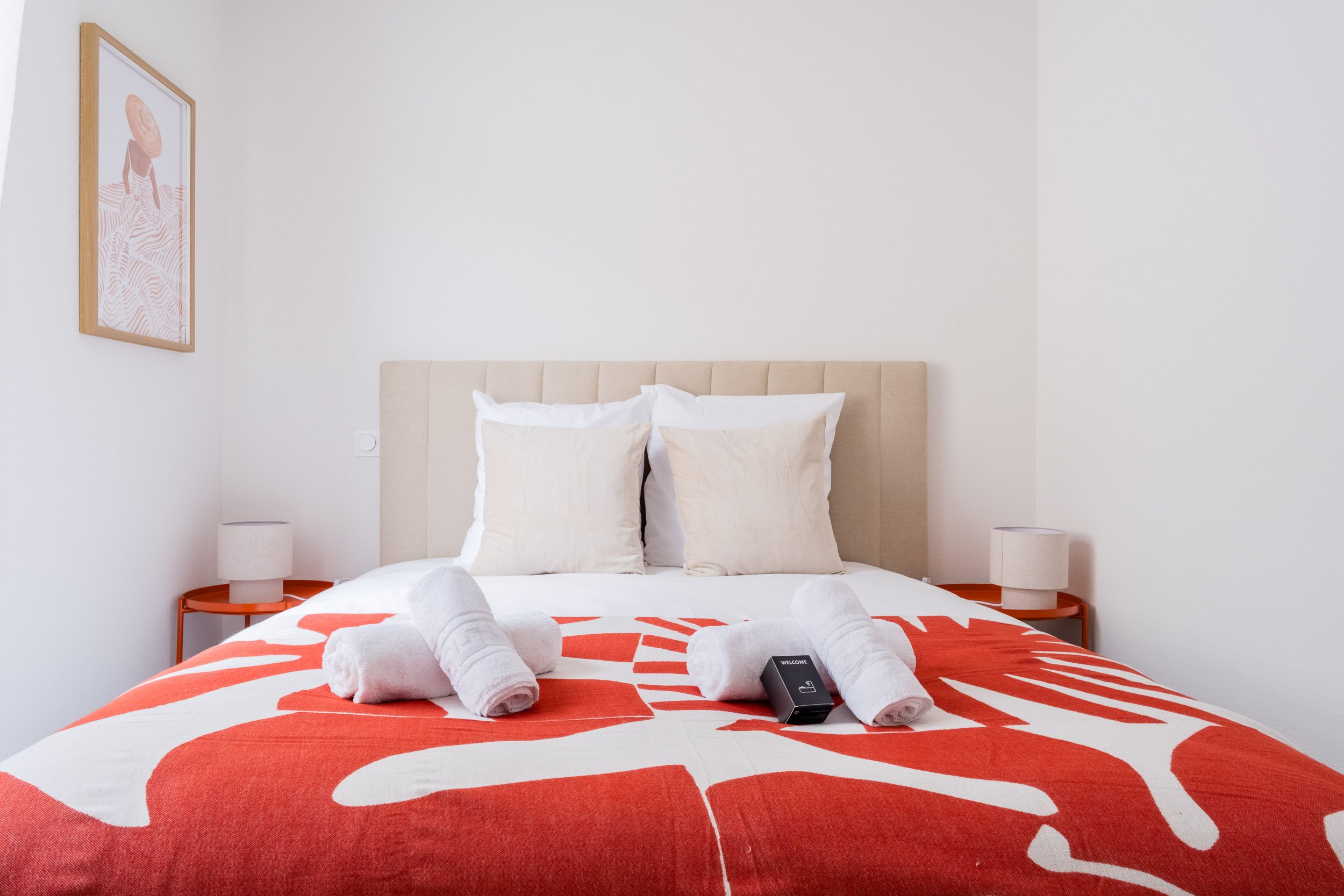 Chambre moderne avec un lit à baldaquin blanc, serviettes roulées sur la couverture rouge à motifs blancs, deux tables de chevet avec lampes, et un tableau d'art sur le mur blanc.
