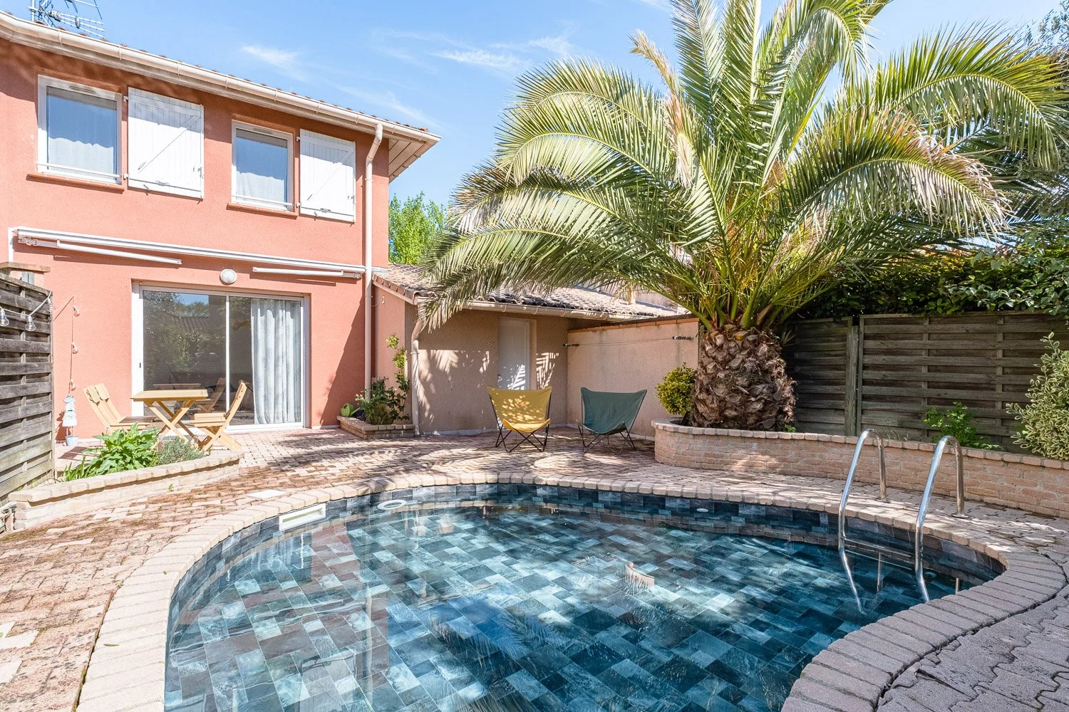 Piscine découverte dans une cour arrière avec une maison rose, des chaises longues, un palmier, et une clôture en bois sous un ciel bleu.