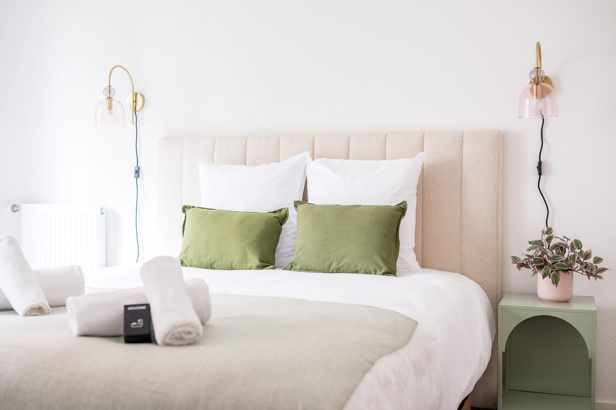 Chambre moderne avec un lit blanc, deux oreillers blancs, deux coussins verts, deux lampes murales en verre rose et laiton, une plante en pot à côté d'une table de nuit verte.