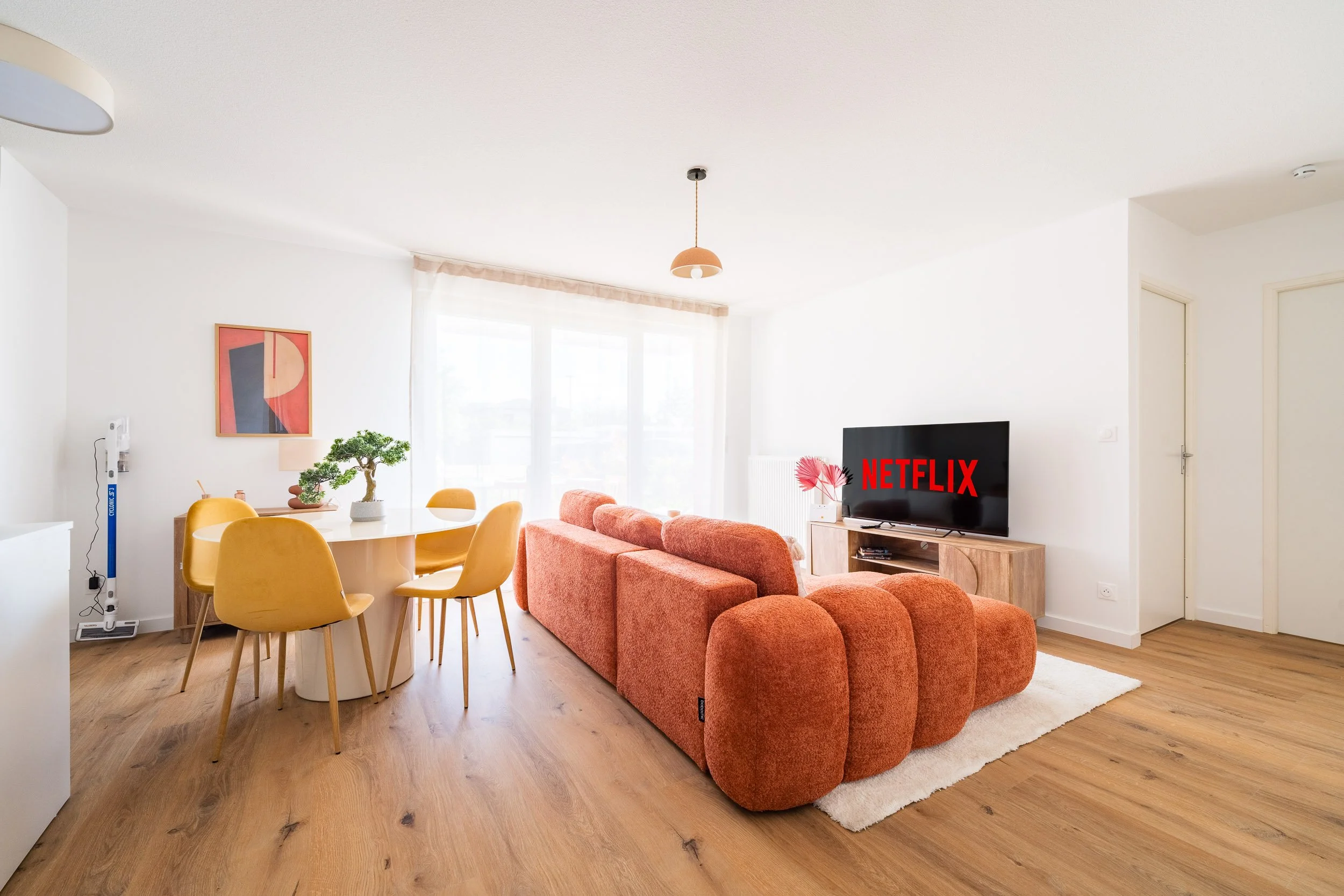 Salon lumineux avec canapé orange, télévision avec le logo Netflix, table ronde avec chaises jaunes, décoration moderne, plantes vertes, et mur blanc avec tableau abstrait.