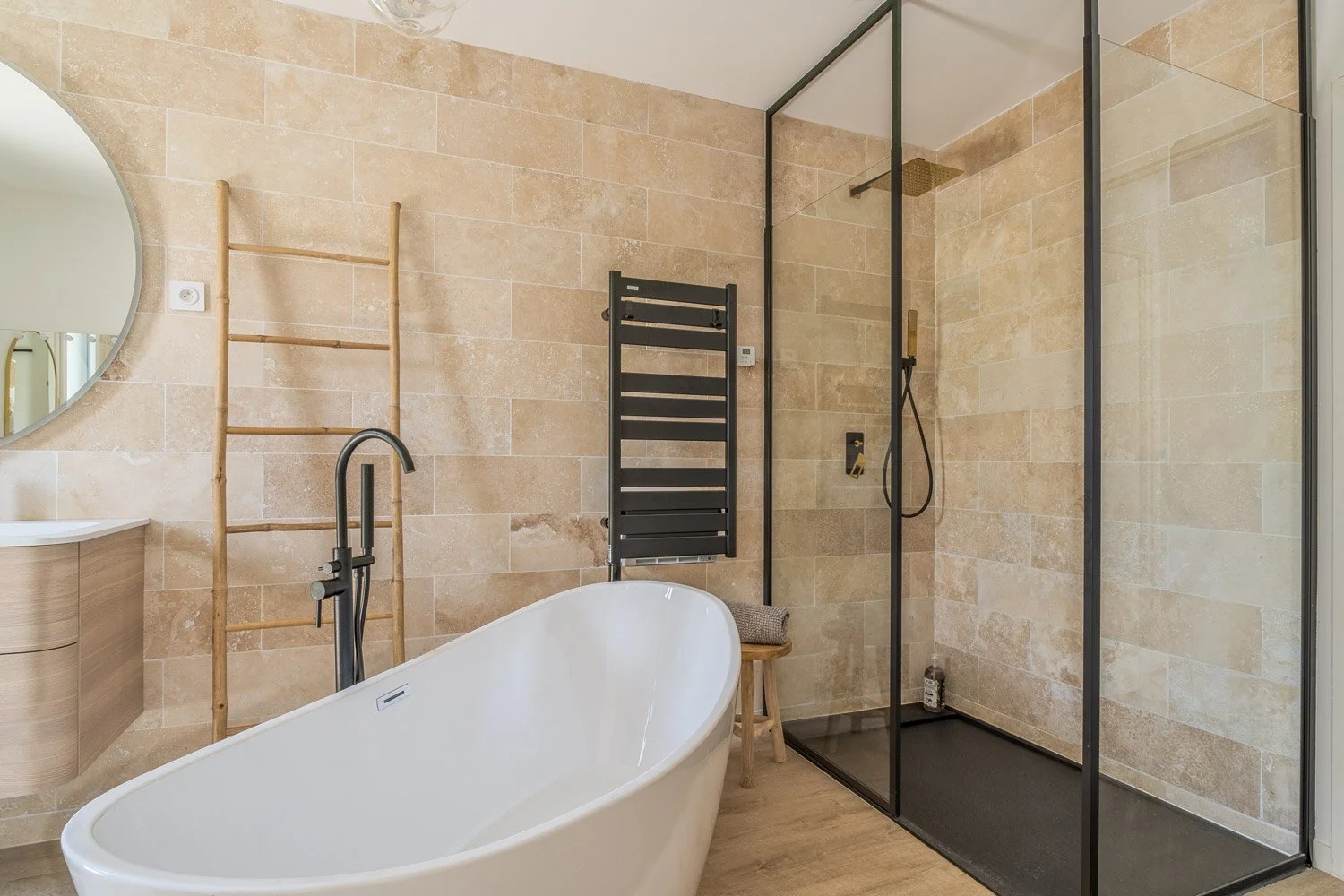 Salle de bain moderne avec une baignoire autoportante, une douche à l'italienne, un lavabo avec meuble en bois, un miroir rond, un porte-serviettes chauffant noir, une échelle en bois pour suspendre des serviettes, et des murs en pierre beige.