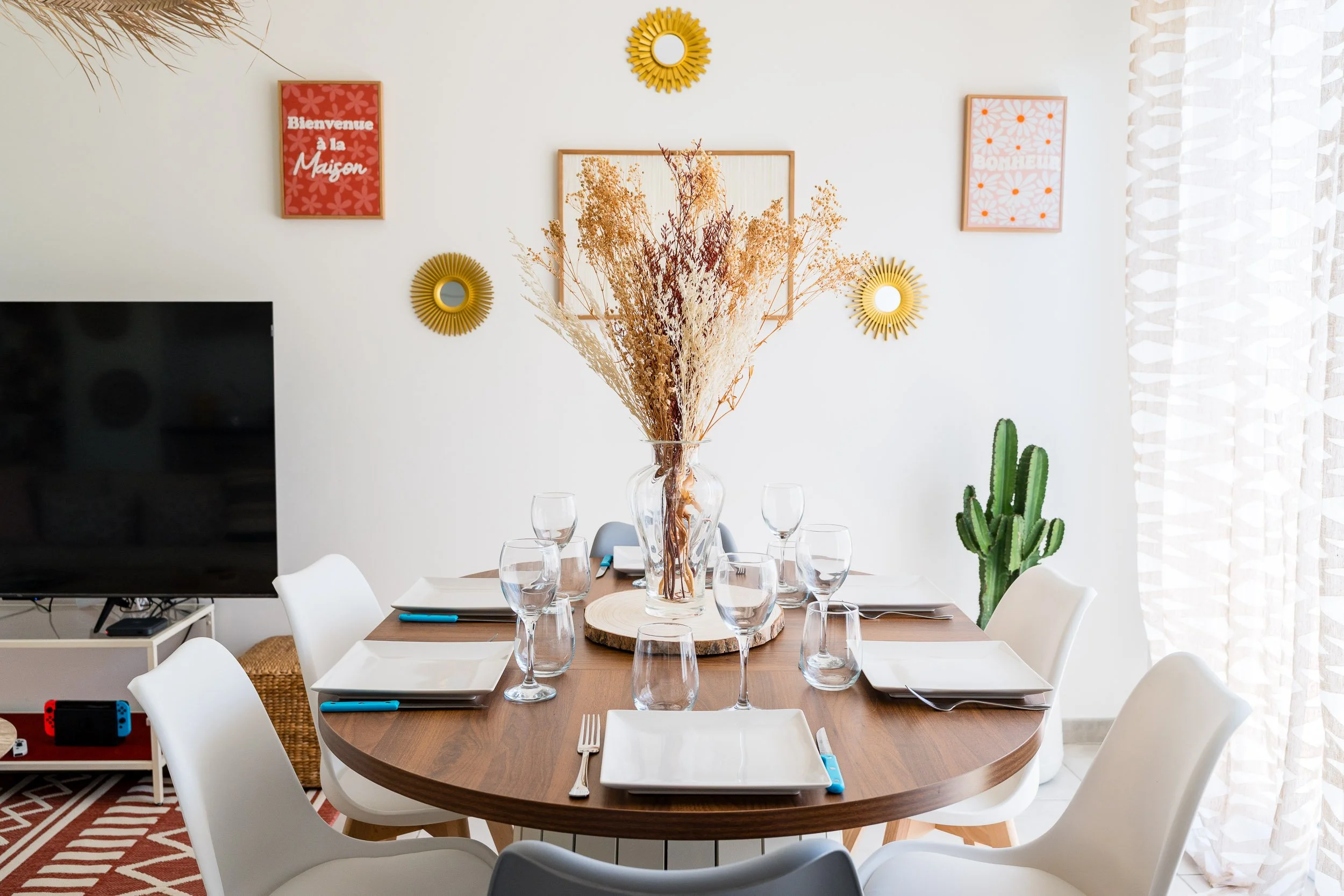 Salle à manger avec un grand vase de branches séchées sur la table, entourée de huit verres et de plaques et couverts. Décor mural comprenant des affiches roses avec des messages en français et des miroirs en forme de soleil. Fenêtres avec rideaux bl