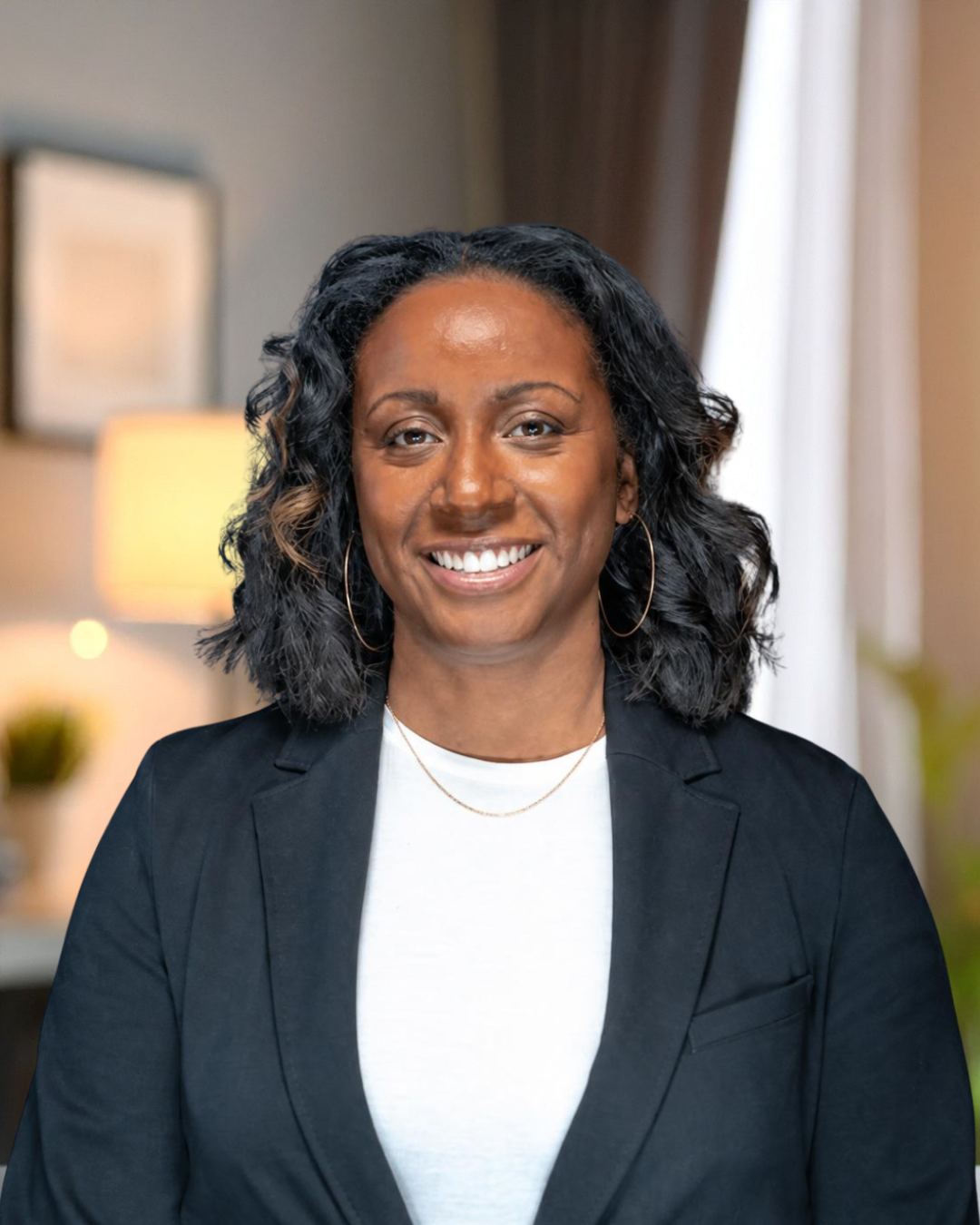 A smiling woman with curly black hair, wearing a black blazer, gold hoop earrings, and a gold necklace, sitting in a cozy, well-lit room.