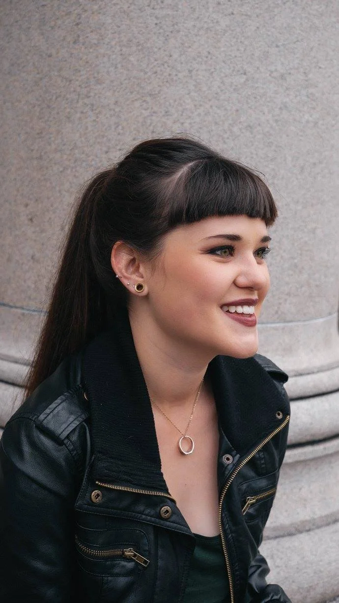 A young woman with dark brown hair and straight bangs, smiling and looking to the right, sitting against a stone wall. She is wearing a black leather jacket, a necklace with a circular pendant, and small earrings.