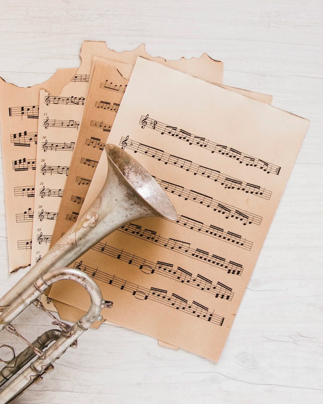 An aged trumpet resting on several sheets of torn and un torn music sheets on a white wooden surface.