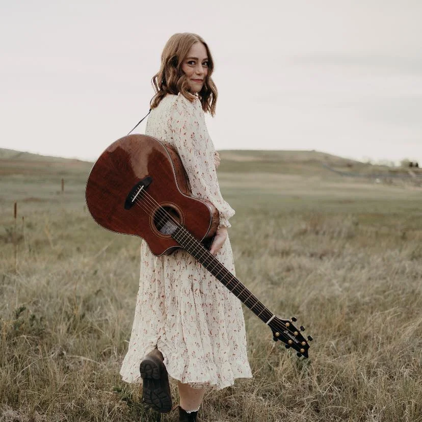 A woman standing in an open grassy field holding an acoustic guitar over her back, wearing a long light-colored dress with a floral pattern and black boots, with a distant horizon and sky in the background.