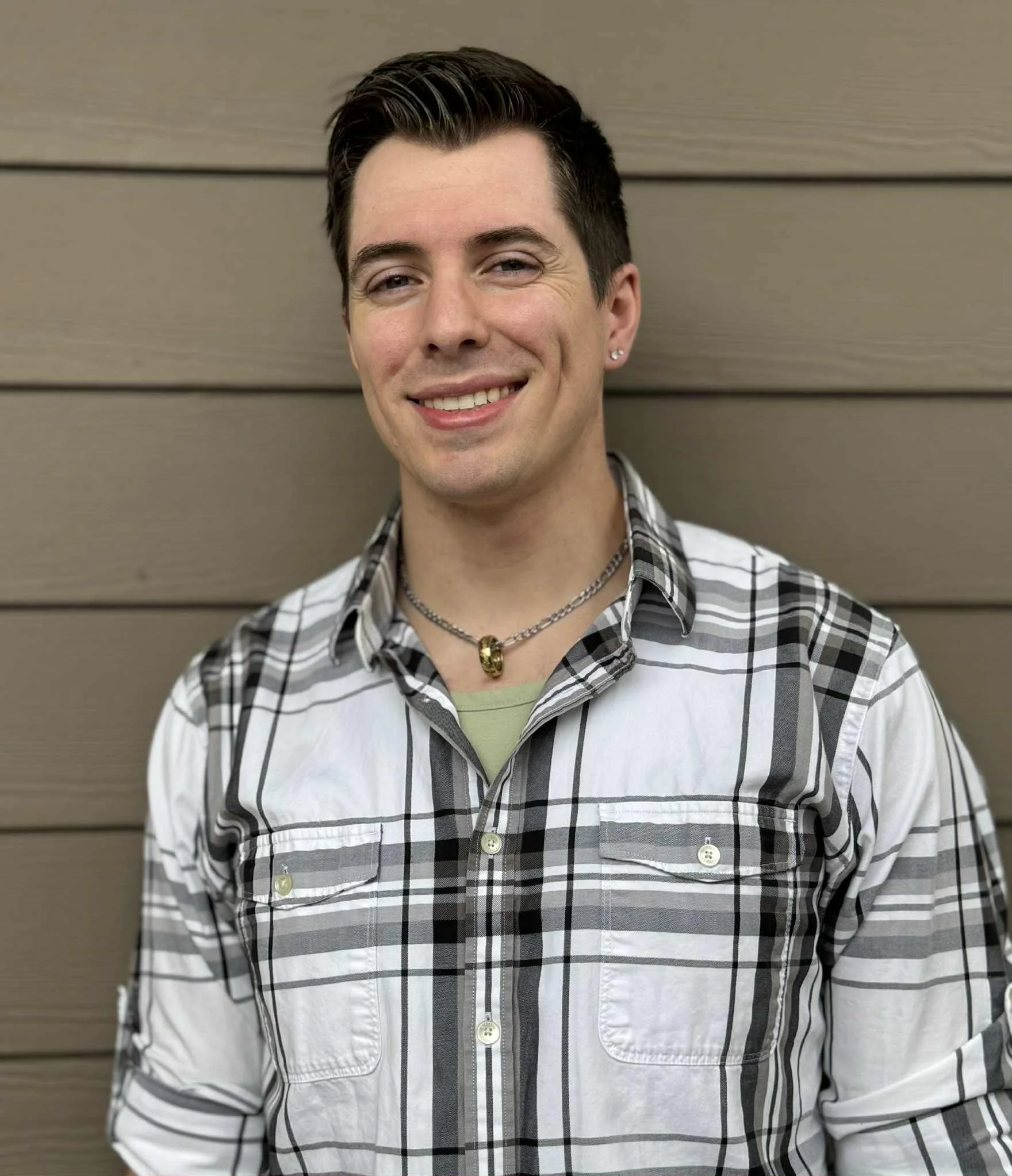 A smiling young man with short dark hair, wearing a plaid button-up shirt, a green undershirt, a necklace, and an earring, standing against a wooden wall.