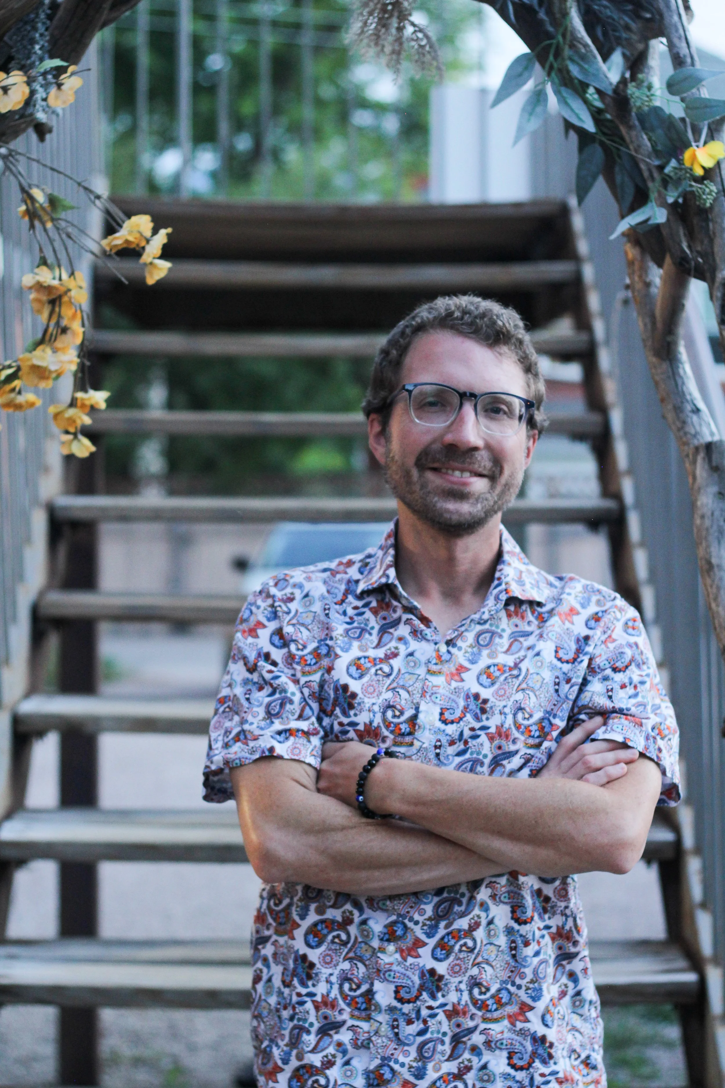 A man with glasses and a beard, smiling with arms crossed, standing in front of a wooden staircase decorated with yellow flowers and greenery.