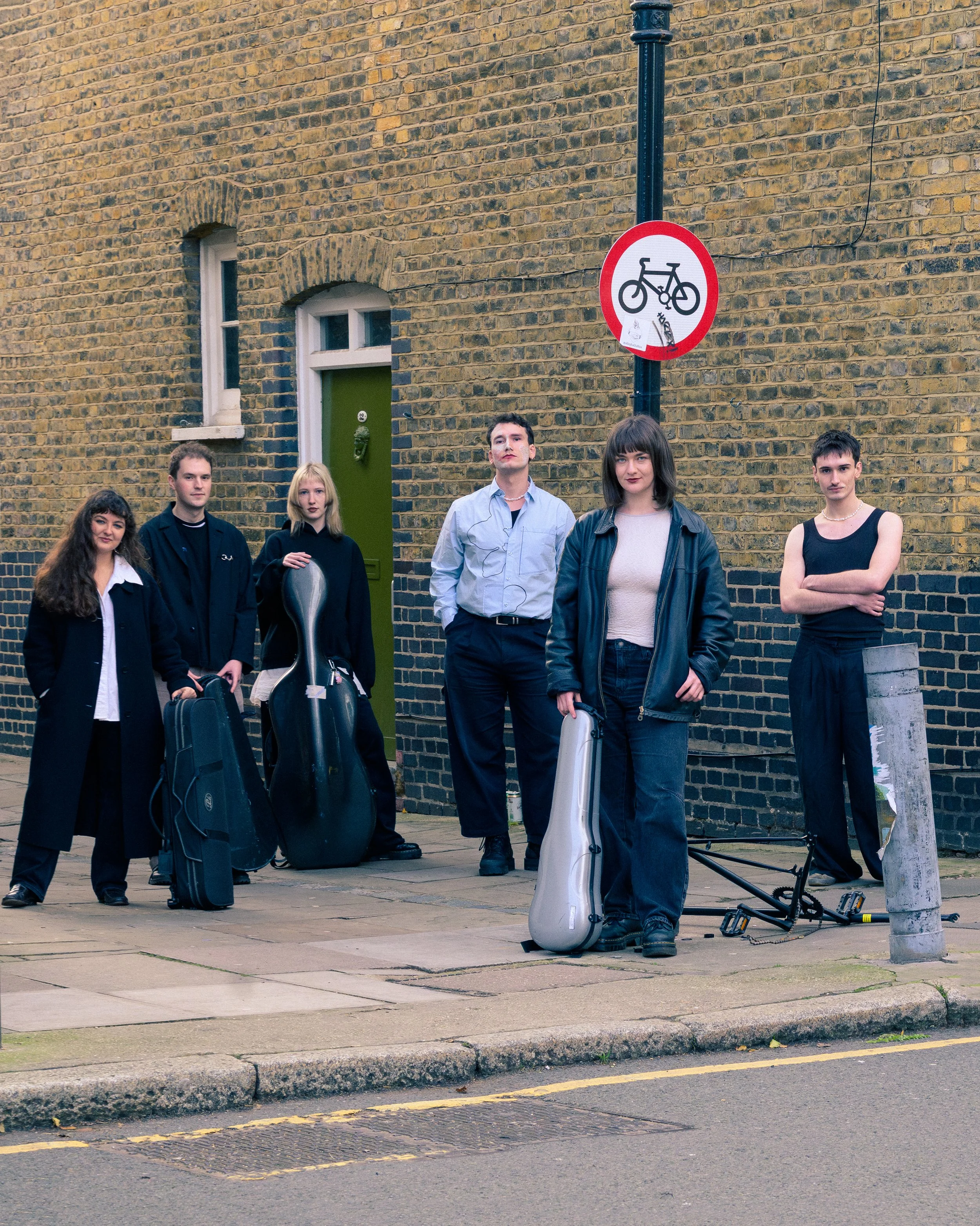Six young adults standing on a sidewalk next to a brick building, some with musical instrument cases, with a no-bicycle sign on a pole behind them.