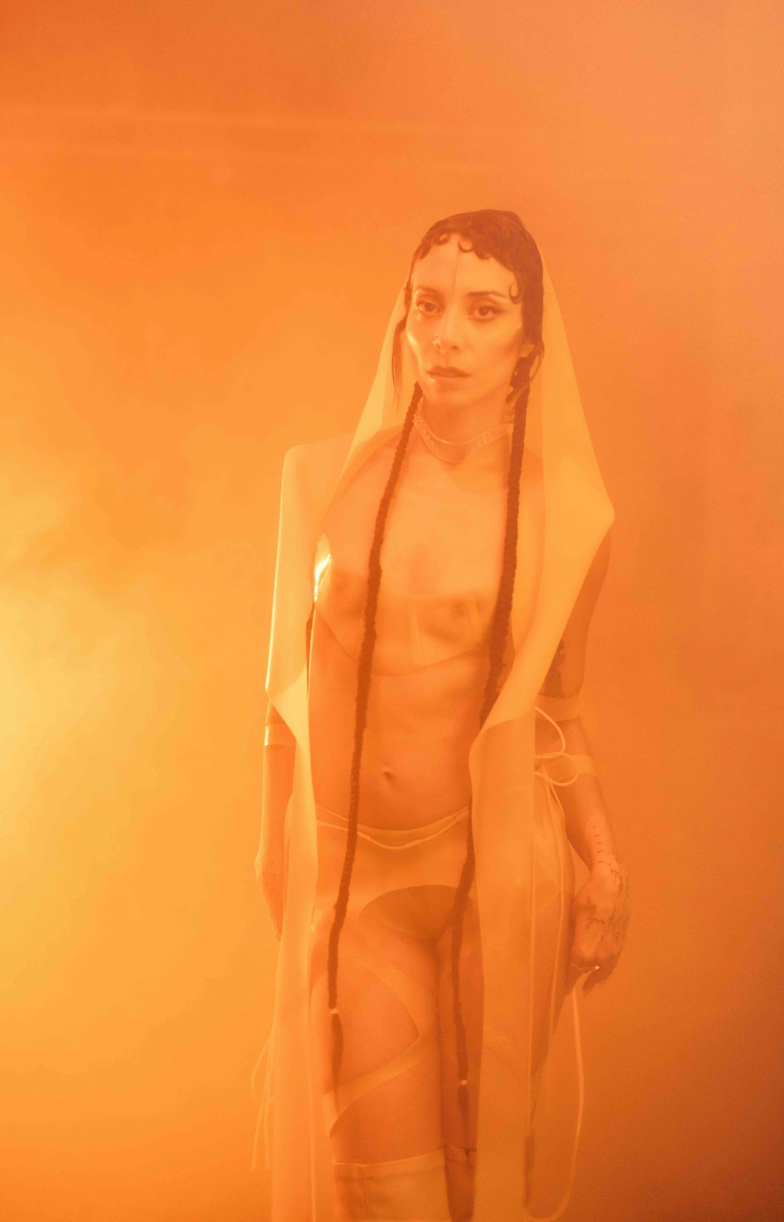 A person with long dark hair, styled in braids, wearing a sheer outfit and a veil, stands in a glowing orange environment.