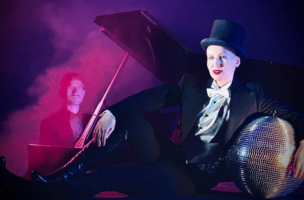 Performer in a tuxedo, bow tie, top hat, and black boots sitting beside a shiny disco ball, with a woman in the background at a piano, all under purple lighting.