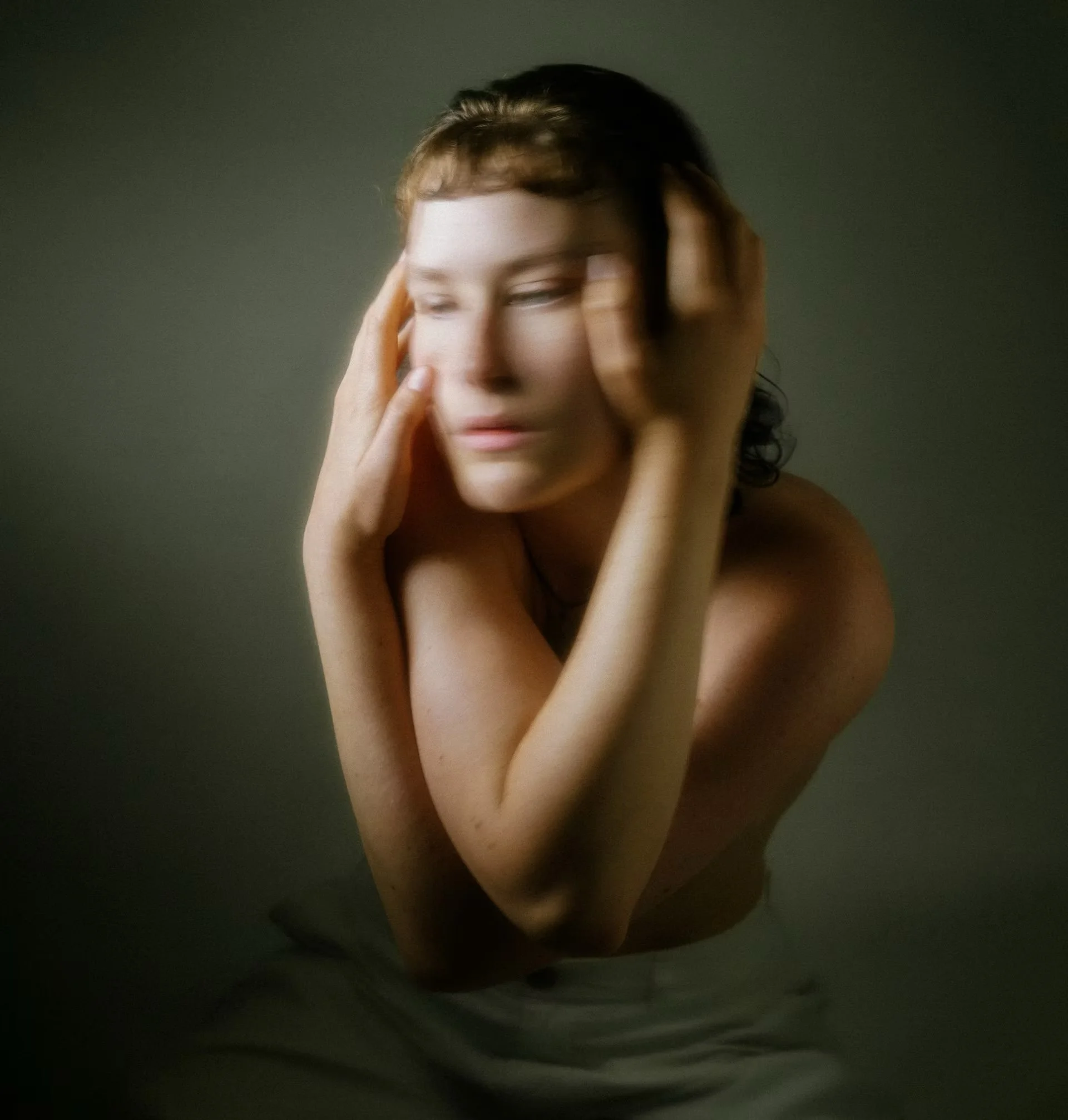 A woman with short dark hair and fair skin is posing with her eyes closed, gently touching her face with both hands against a dark background.