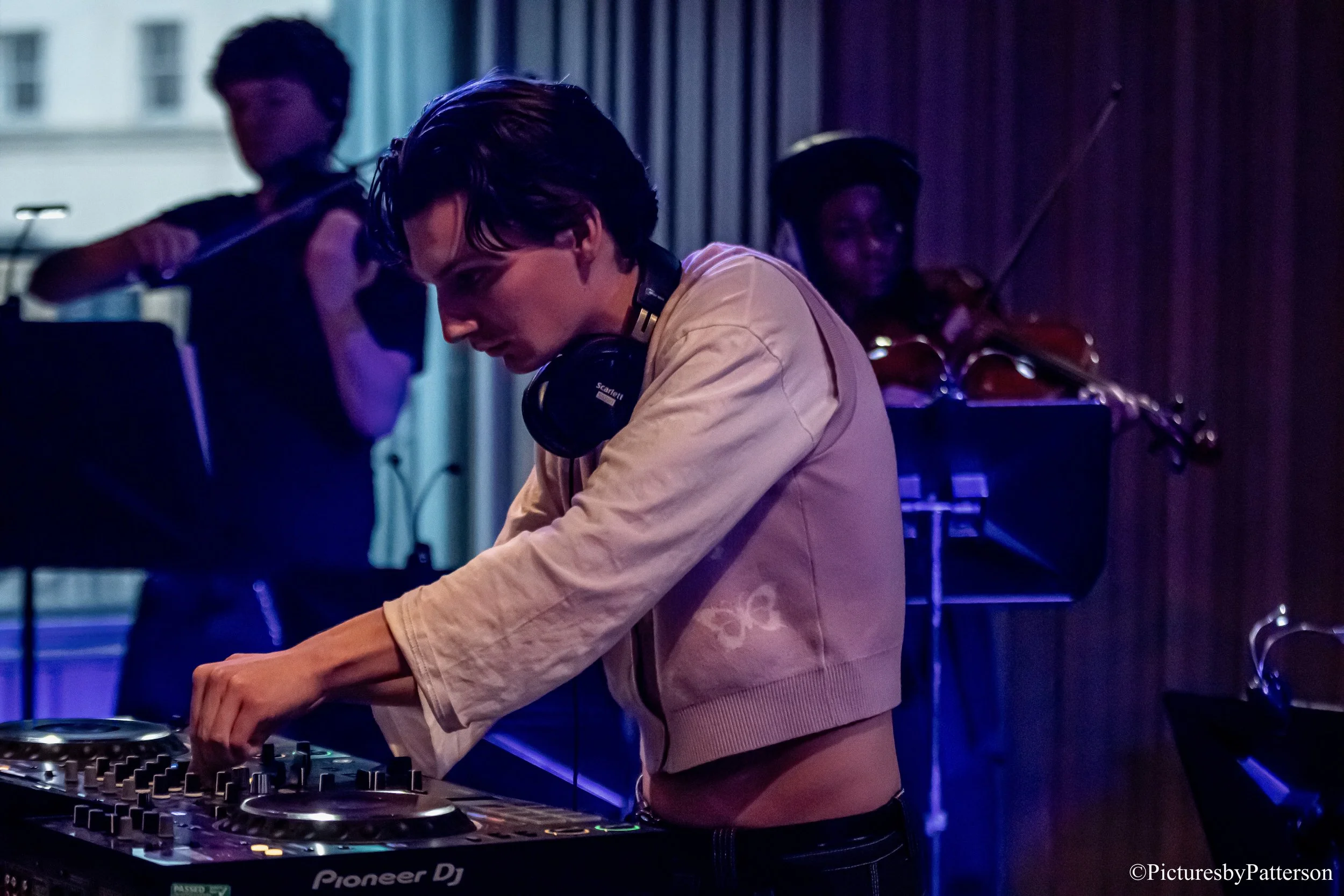 A DJ wearing a beige jacket and headphones operating a Pioneer DJ controller, with two violin players in the background in a dimly lit room.